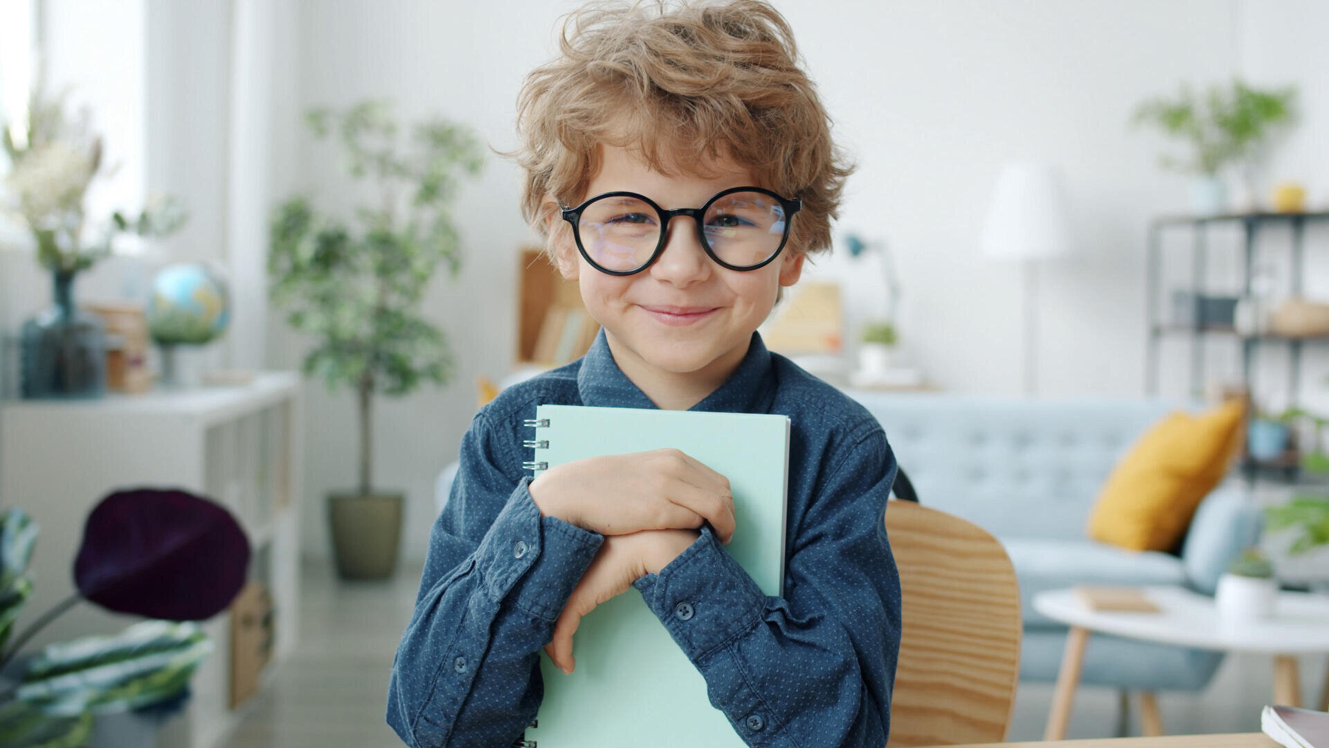 A smiling young boy with curly hair and large round glasses hugs a light blue notebook while sitting at a desk in a bright, cozy room with plants and modern decor.