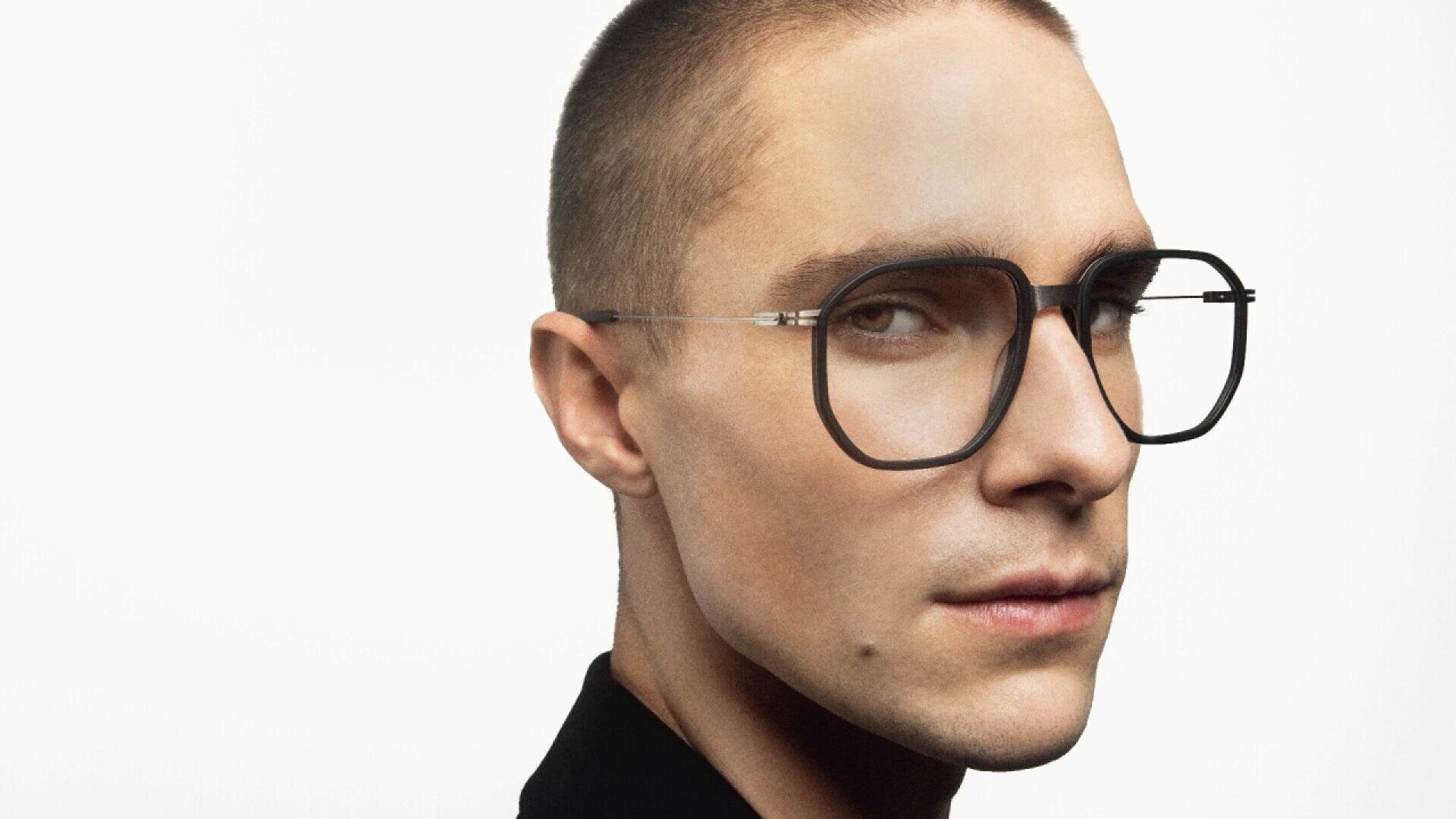 Eine Person mit kurzen Haaren und einer großen, geometrischen schwarzen Brille blickt vor einem schlichten weißen Hintergrund leicht zur Seite und trägt ein dunkles Oberteil.