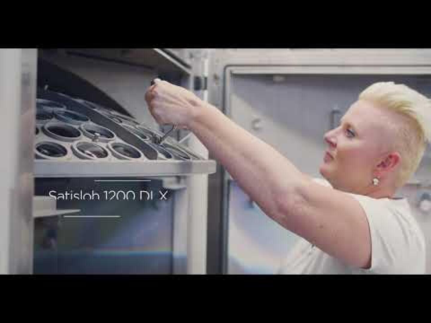 Eine Person mit kurzen blonden Haaren bedient eine Maschine und setzt runde Metallschalen ein oder entnimmt sie. Der Text Satisloh 1200 DLX erscheint auf dem Bild. Die Umgebung sieht aus wie eine Industrie- oder Laborumgebung.