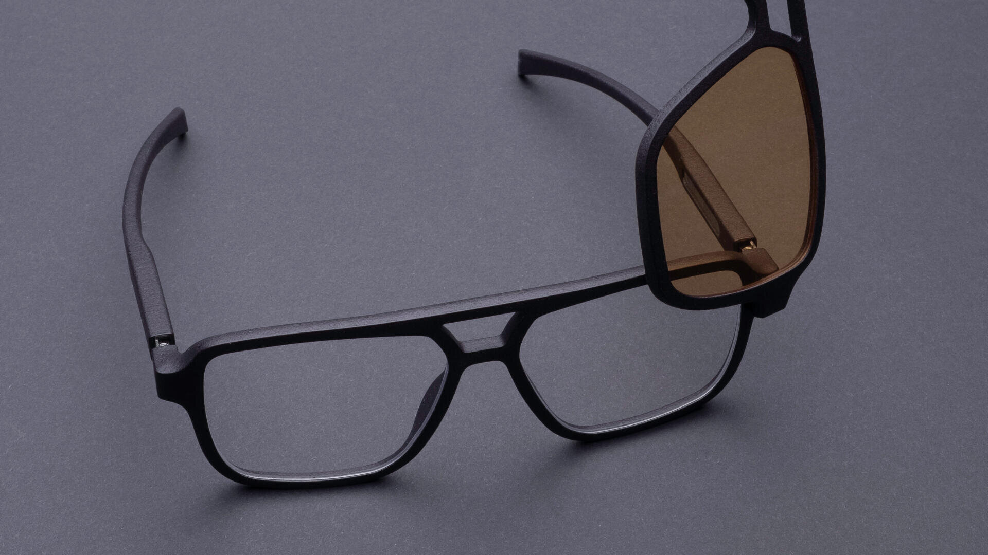 Zwei Brillen liegen auf einer dunkelgrauen Fläche. Eine hat klare Gläser, die andere getönte braune Gläser. Die Brillen sind so angeordnet, dass sich ihre Rahmen vorne überlappen.