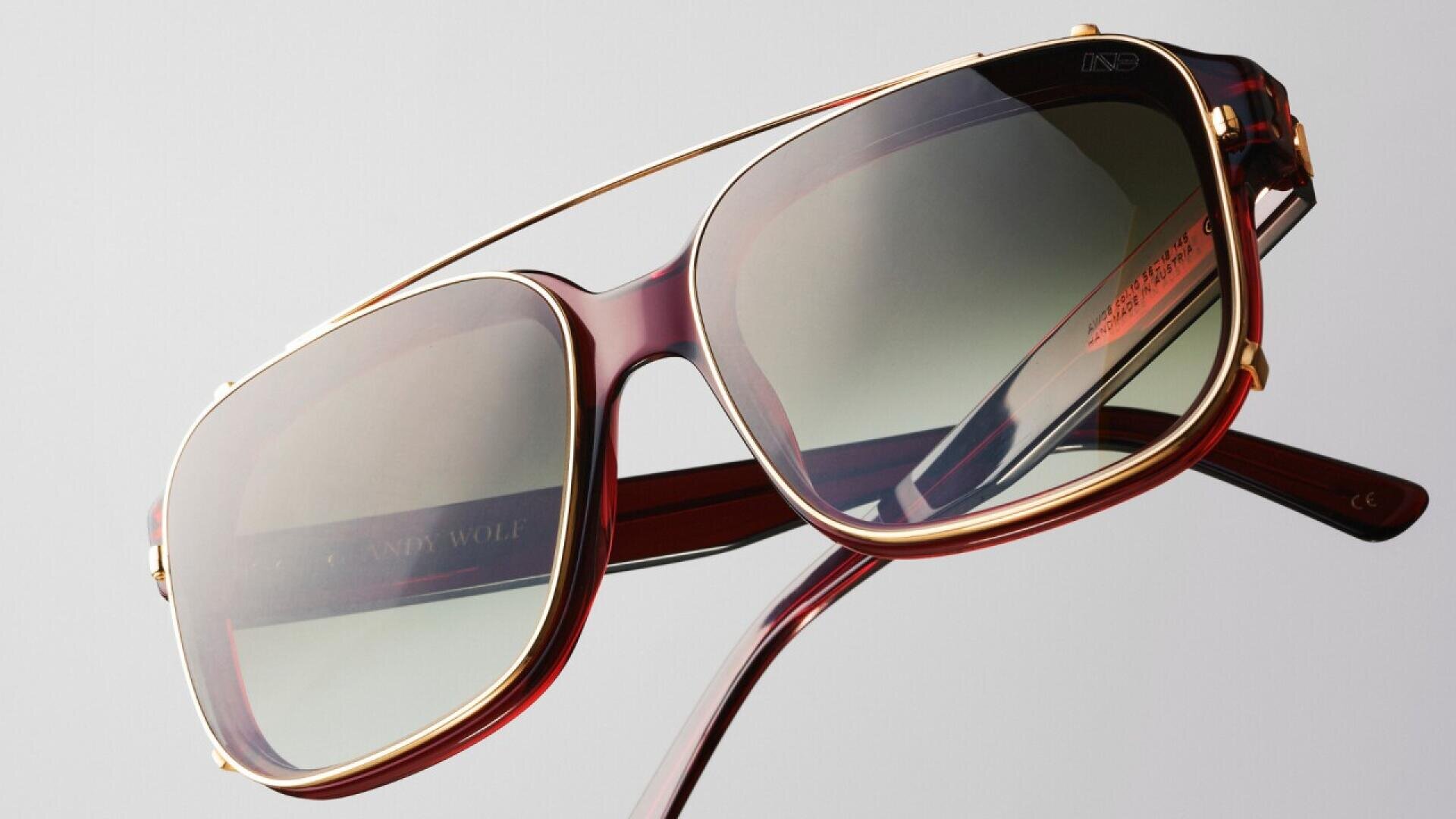 Close-up of stylish sunglasses with a maroon and gold frame, green tinted lenses, and a thin gold brow bar, set against a plain light background.