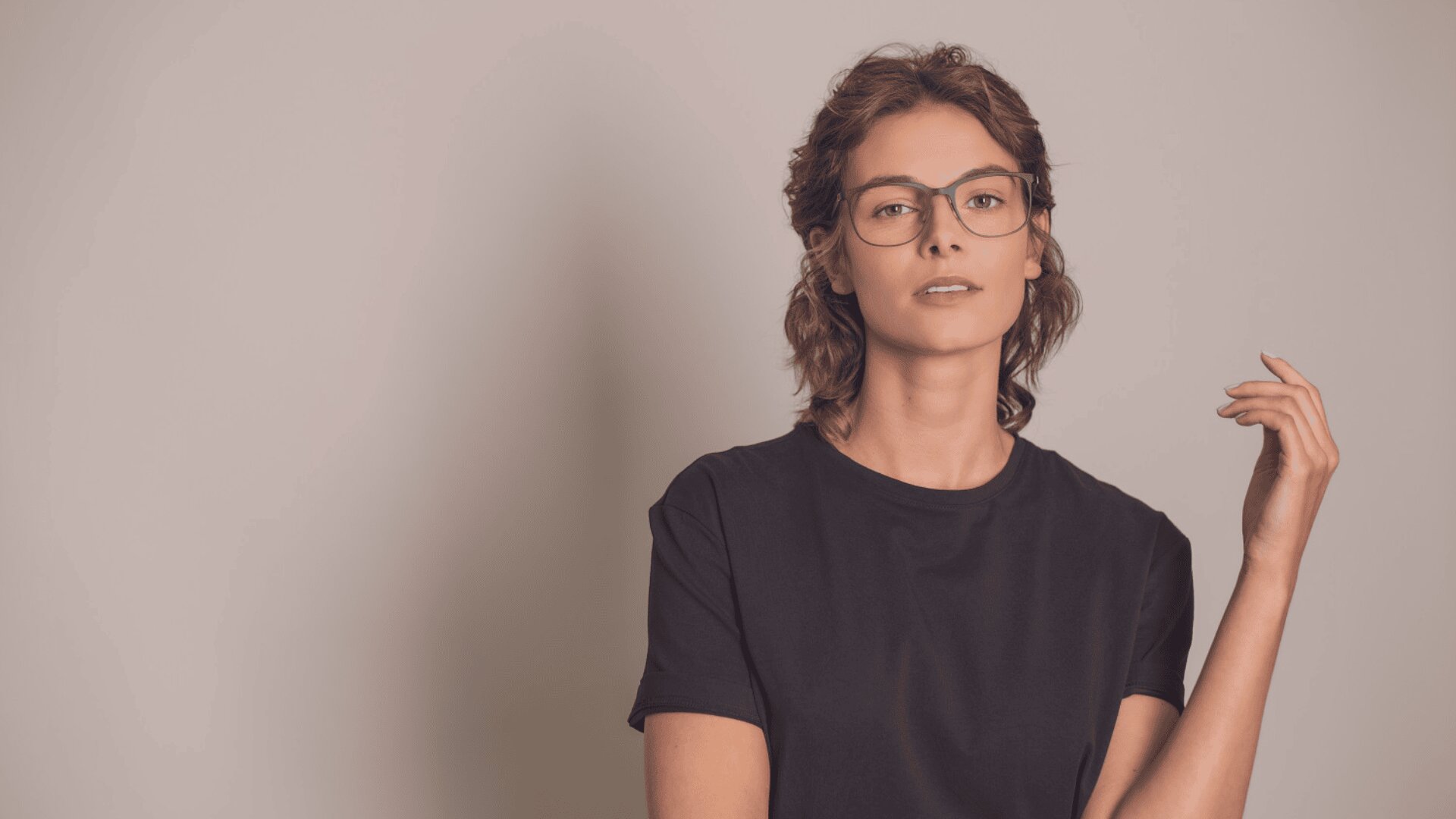 A person with short, wavy brown hair wearing glasses and a plain black t-shirt stands against a light beige background, looking at the camera with a neutral expression and one hand raised.