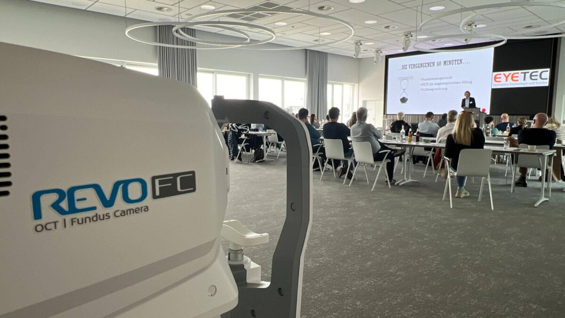 Im Vordergrund eine Nahaufnahme einer REVO FC OCT/Funduskamera, im Hintergrund eine Gruppe von Menschen, die an Tischen sitzen und einer Präsentation in einem modernen Konferenzraum zuhören.