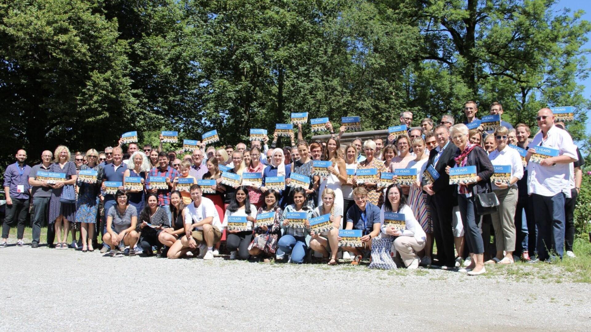 A large group of people stand outdoors on a sunny day, smiling and holding rectangular signs with blue backgrounds. Trees and greenery are visible in the background.