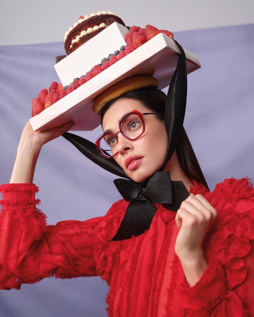 A woman in a bright red textured dress and red glasses balances a tray with a cake and berries on her head, secured with a large black ribbon. She poses against a light purple background.