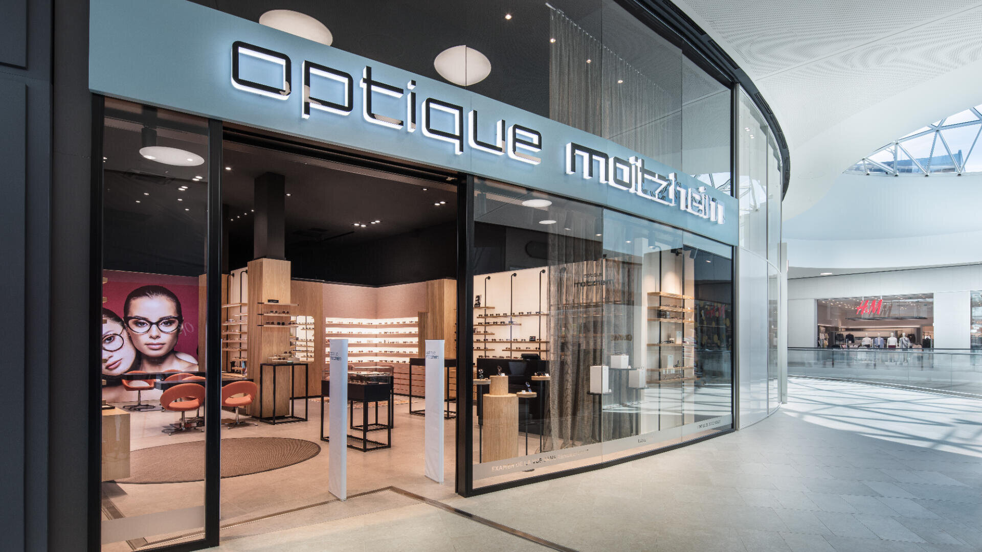 Ein modernes Optikergeschäft namens Optique Moitzheim mit stilvoller Glasfassade, Brillendisplays und Sitzgelegenheiten im Inneren. Ein großes Poster mit Gesichtern mit Brillen ist zu sehen. Draußen sind das Innere des Einkaufszentrums und eine H&M-Filiale zu sehen.