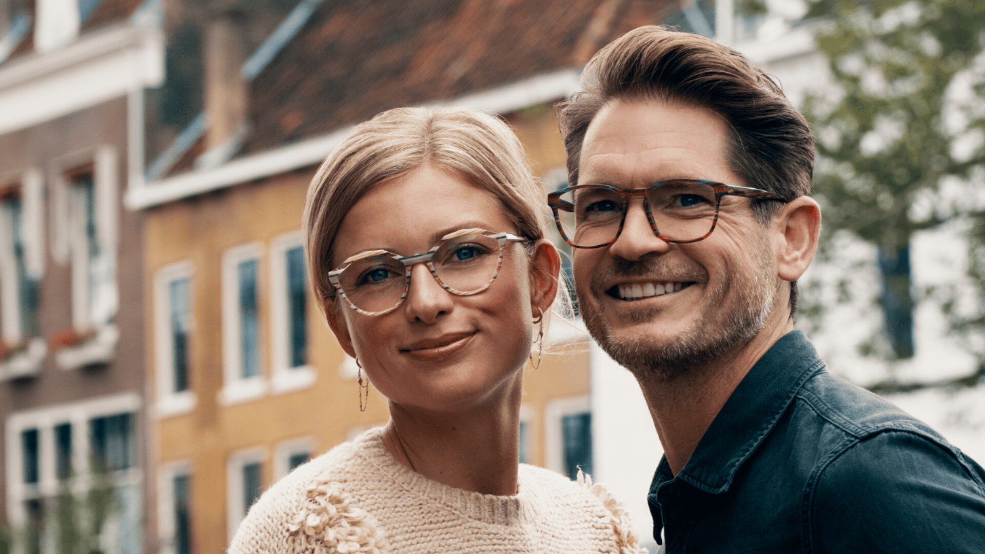 Eine lächelnde Frau und ein Mann mit Brille stehen dicht beieinander im Freien, mit bunten Gebäuden und Grün im unscharfen Hintergrund.