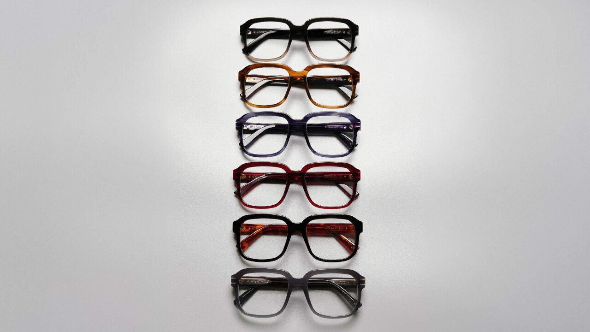Six pairs of eyeglasses with thick frames in various colors (black, brown, blue, red, orange, and dark purple) are arranged vertically on a light gray background.