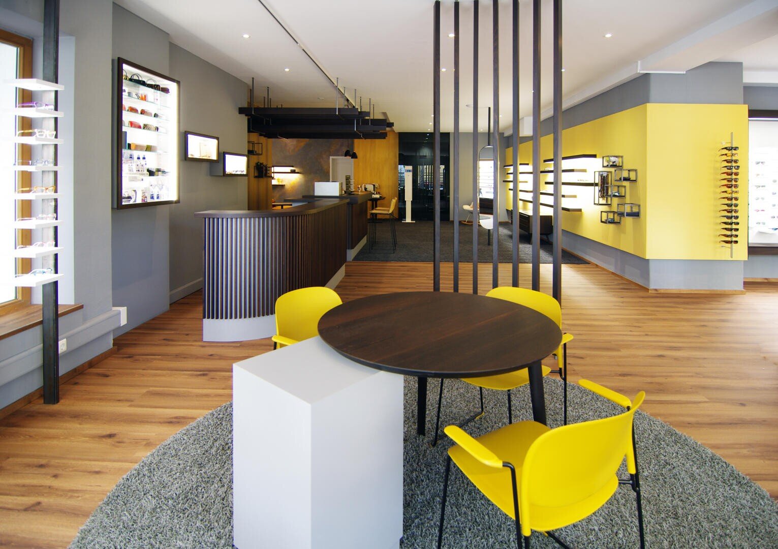 A modern optician's store with yellow chairs around a round table, wooden floors, gray and yellow walls, eyeglass displays and large windows that let in natural light.