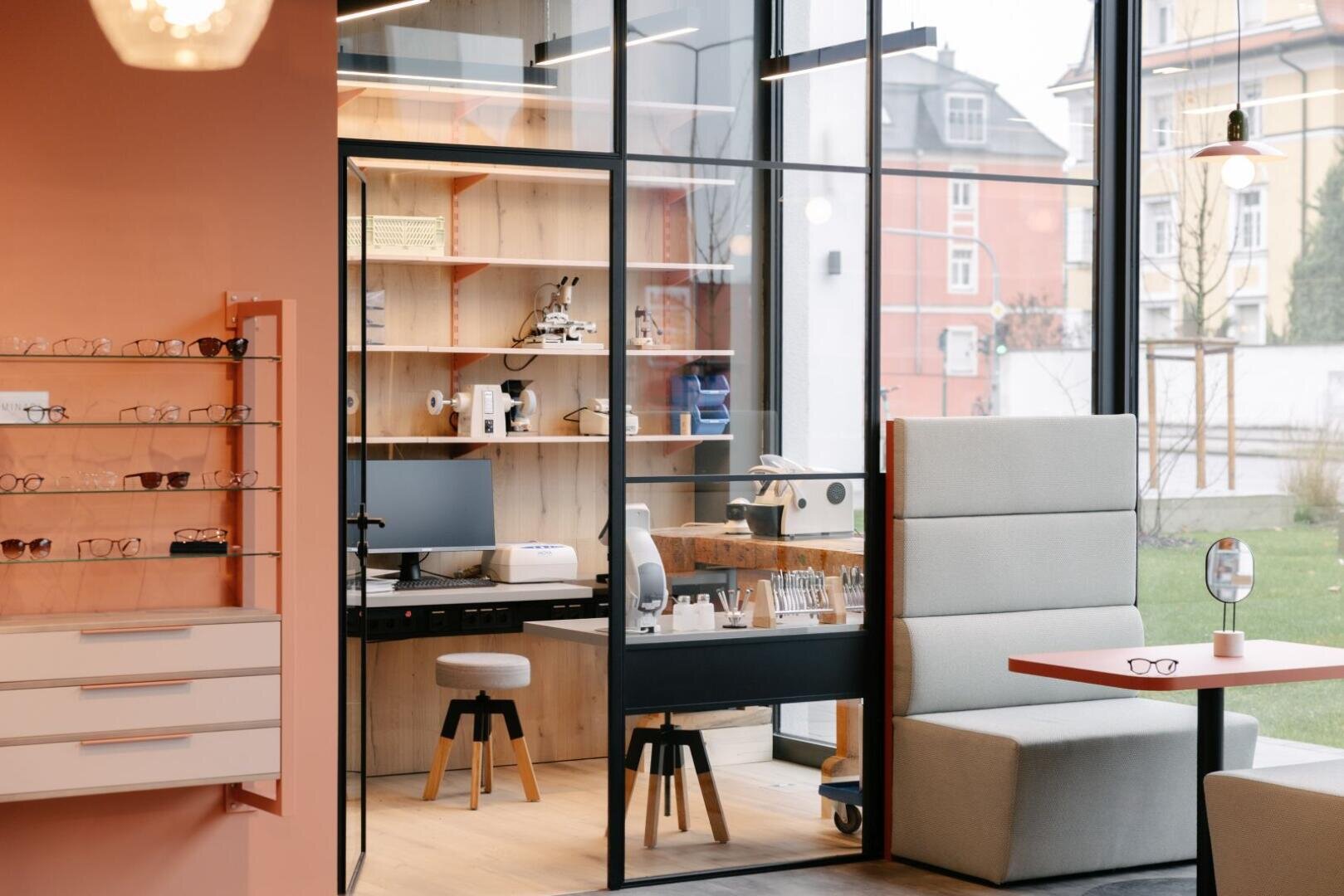 Modernes Optikergeschäft mit großen Fenstern, einem durch Glaswände abgetrennten Untersuchungsraum, Regalen mit Brillengestellen, einem Schreibtisch mit optischen Geräten und gemütlichen Sitzgelegenheiten. Draußen sind durch die Fenster bunte Gebäude zu sehen.