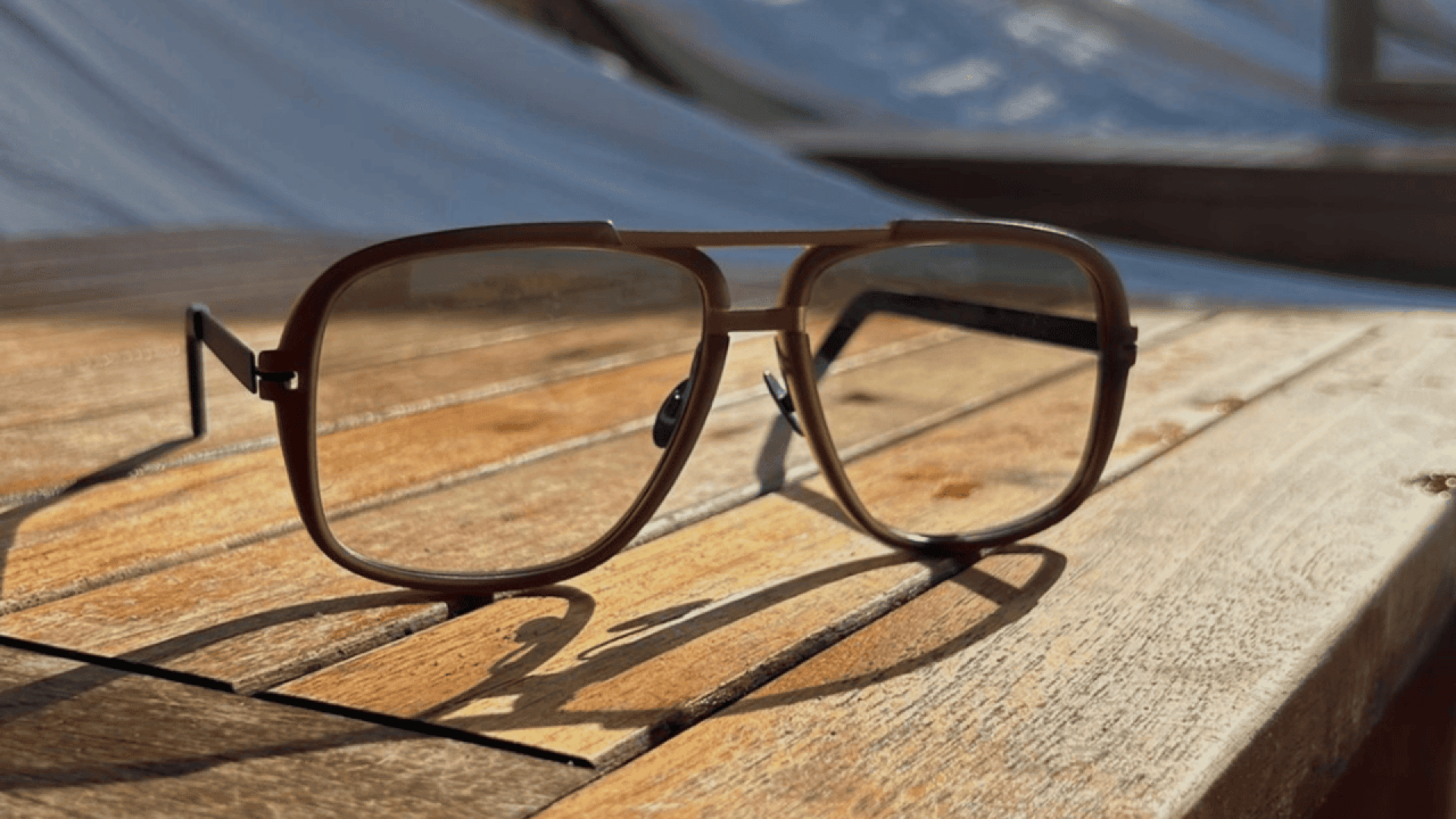 A pair of brown-framed aviator eyeglasses rests on a wooden table in sunlight, casting a clear shadow. The background is blurred, with hints of outdoor furniture and white fabric.