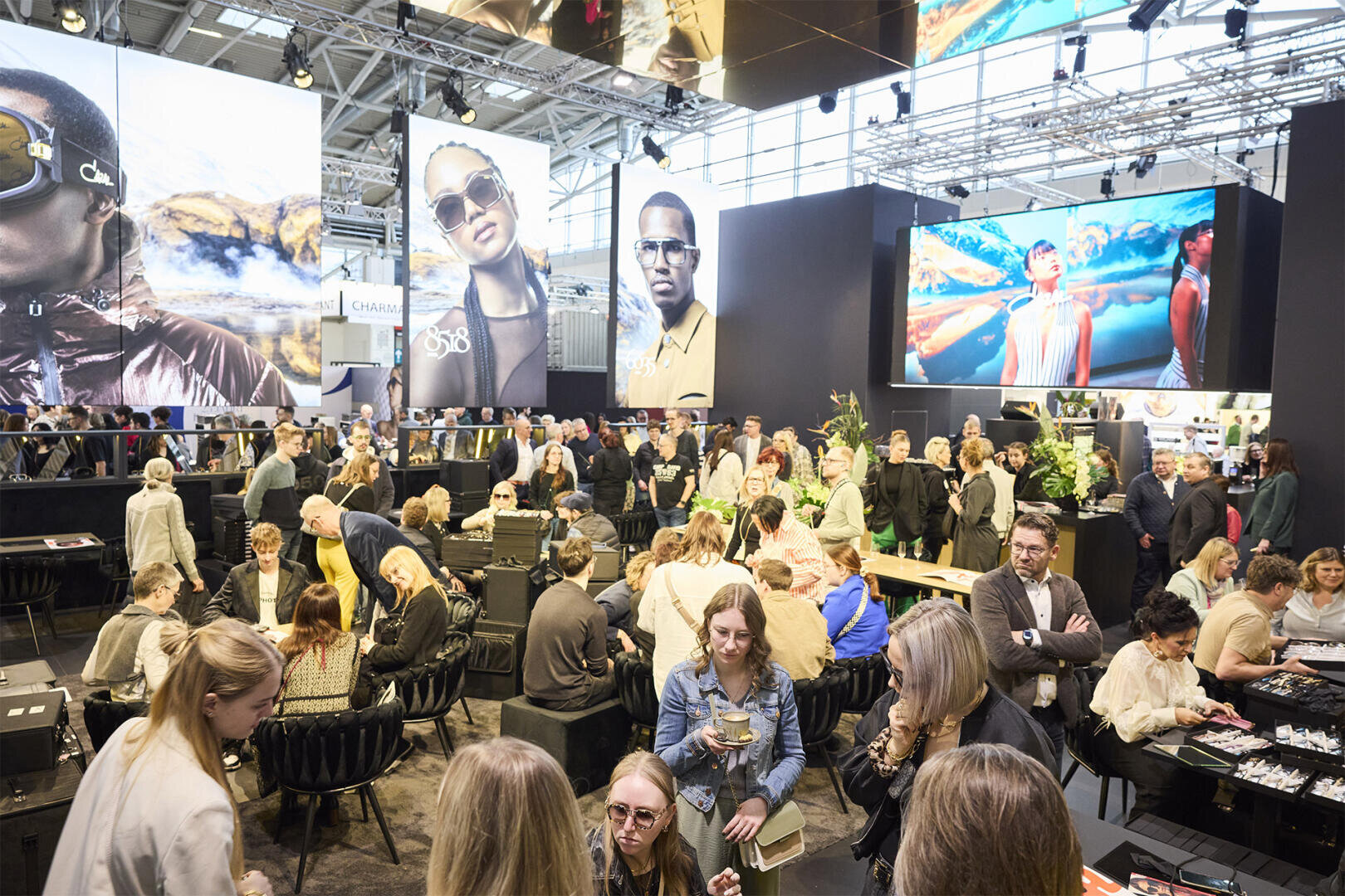 Eine große Gruppe von Menschen versammelt sich bei einer Indoor-Veranstaltung mit Tischen und Stühlen, umgeben von Pflanzen und großen Werbeplakaten, auf denen Models mit Sonnenbrillen abgebildet sind. Helle Beleuchtung erhellt den geräumigen, modernen Veranstaltungsort.