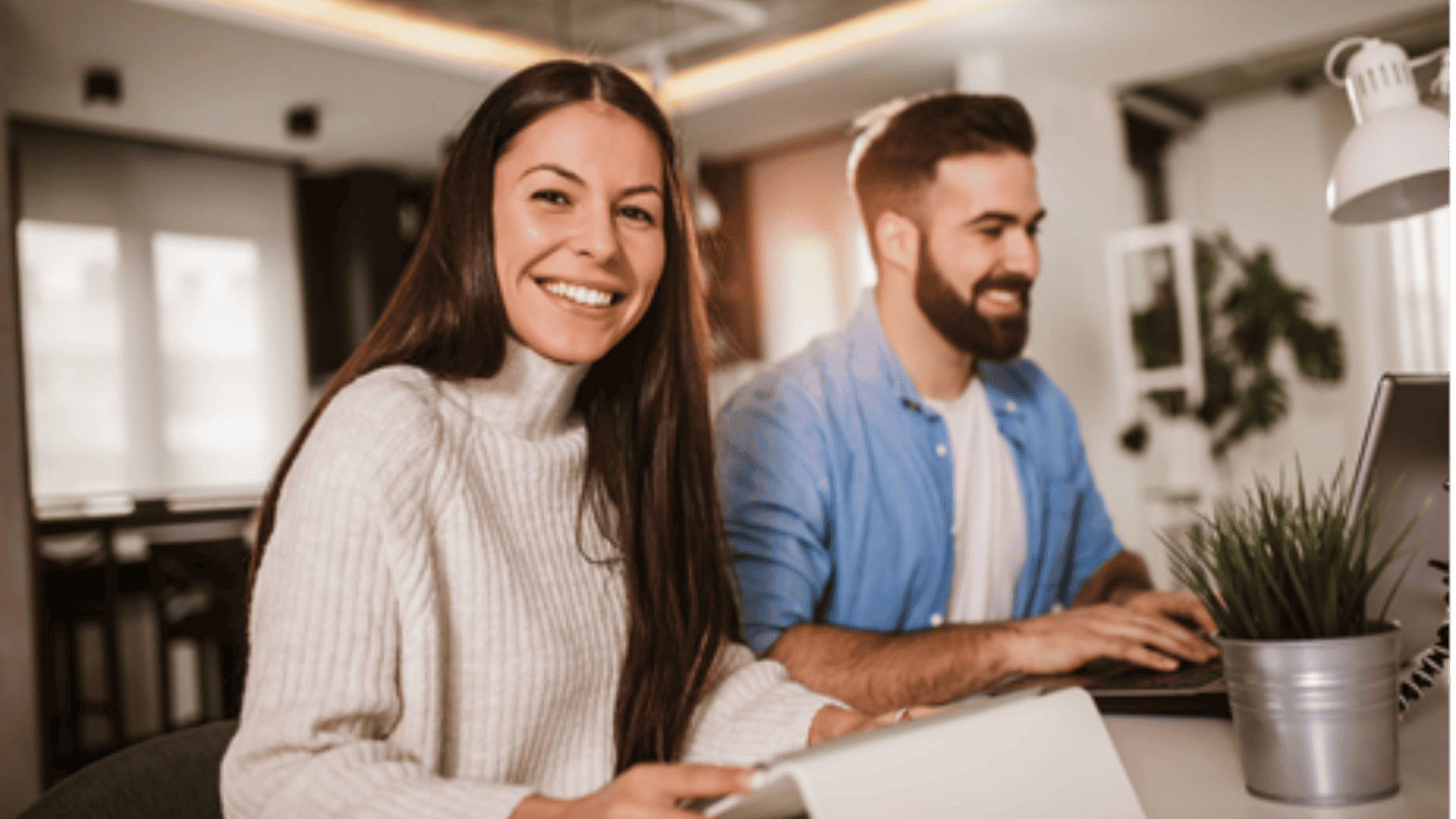 A woman in a white sweater smiles at the camera while sitting at a desk with a laptop. Next to her, a man in a blue shirt is working on a laptop. They are in a modern, well-lit office.