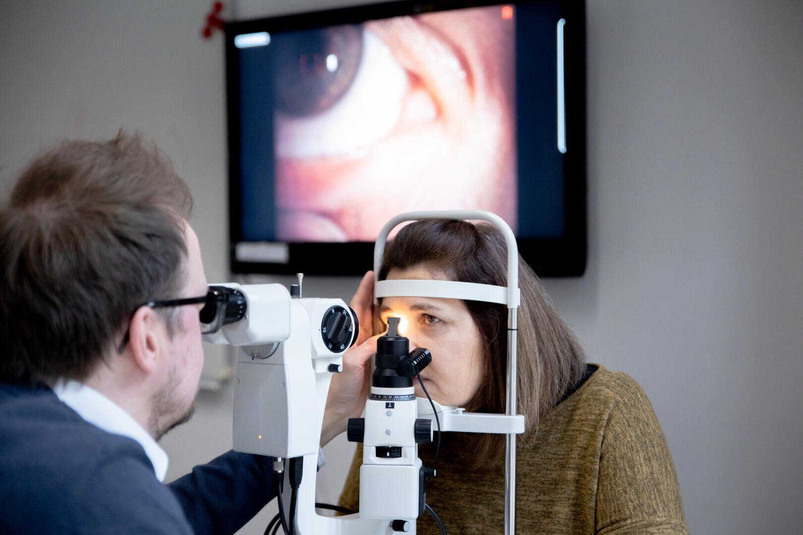 A woman undergoes an eye exam with a slit lamp while a doctor observes; an enlarged image of her eye is displayed on a screen in the background.