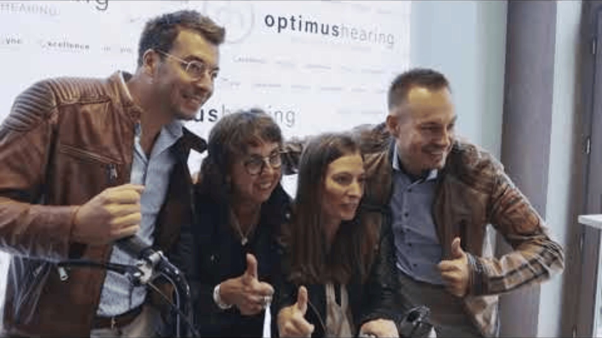 Four people stand close together, smiling and giving thumbs up in front of an Optimus Hearing banner. They appear to be celebrating or posing for a group photo at an event.