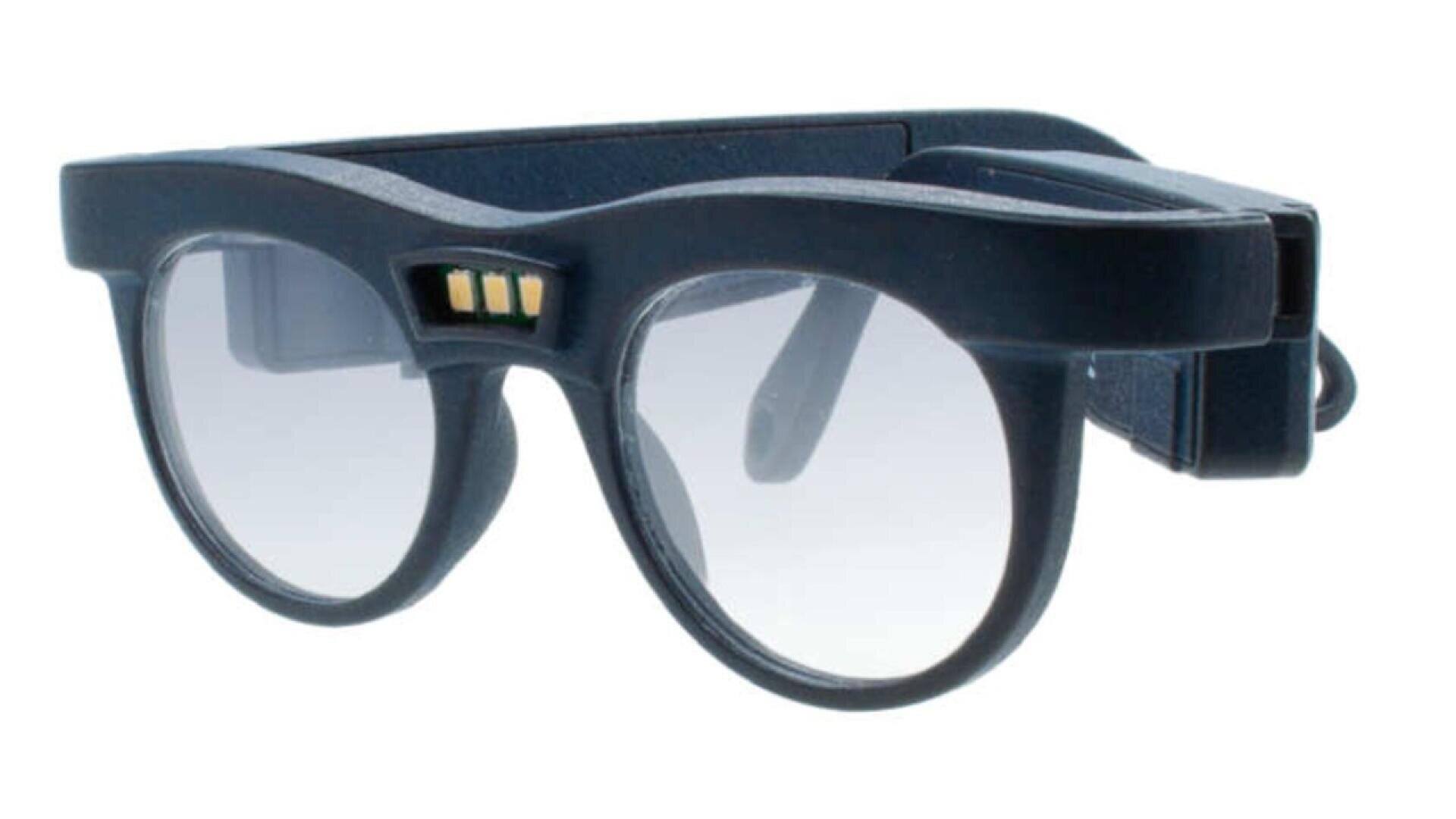A pair of black smart glasses with thick frames, clear lenses, and gold sensor contacts on the bridge. The arms are bulky, indicating built-in electronic components.