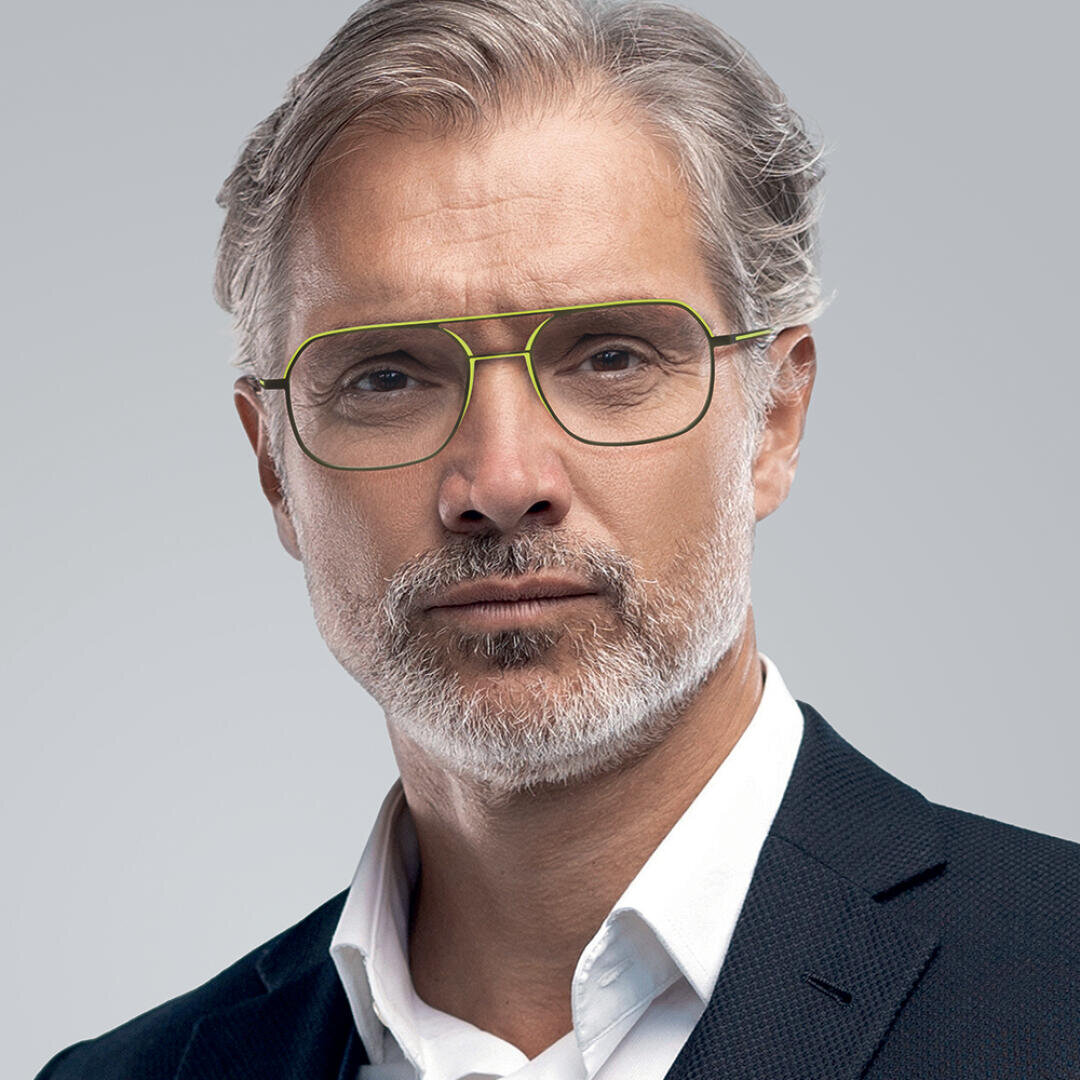 Ein Mann mittleren Alters mit grauem Haar, einem ordentlich gestutzten Bart und Schnurrbart trägt eine grün umrandete Brille und ein dunkles Jackett über einem weißen Hemd und posiert vor einem hellgrauen Hintergrund.