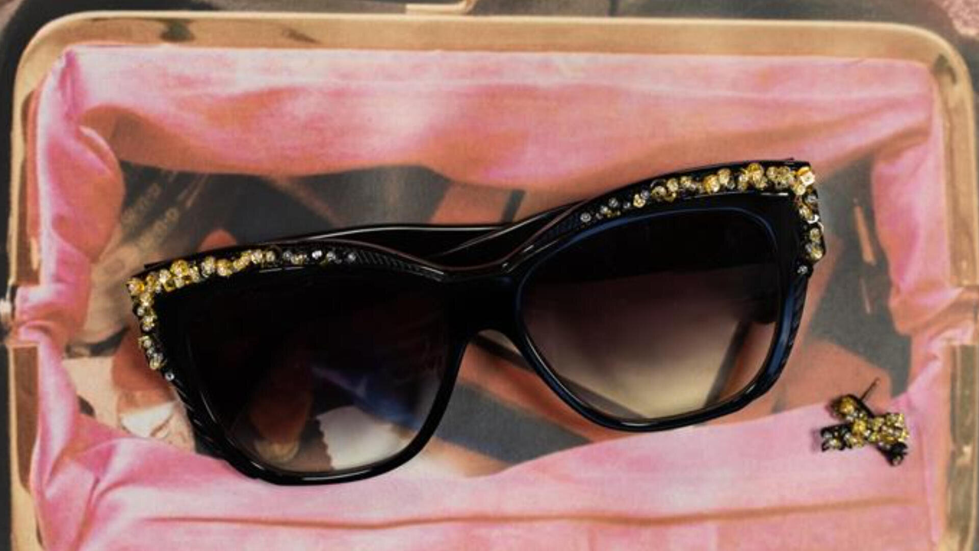 Eine schwarze Cat-Eye-Sonnenbrille mit goldenen Strasssteinen am oberen Rand liegt auf einer rosa Stoffoberfläche neben einer kleinen, mit Schmucksteinen besetzten Bienenbrosche.
