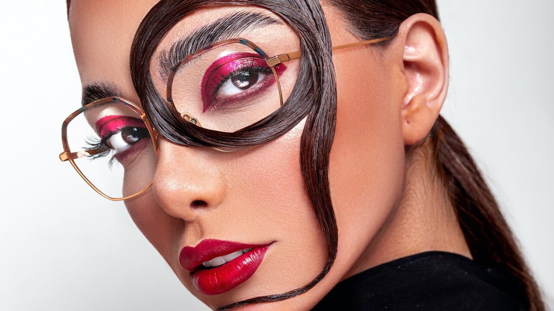 A woman wearing gold-rimmed glasses has sleek dark hair styled in a swirl across her face. She has bold red eyeshadow and lipstick, and her side-parted hair is pulled back. The background is white.