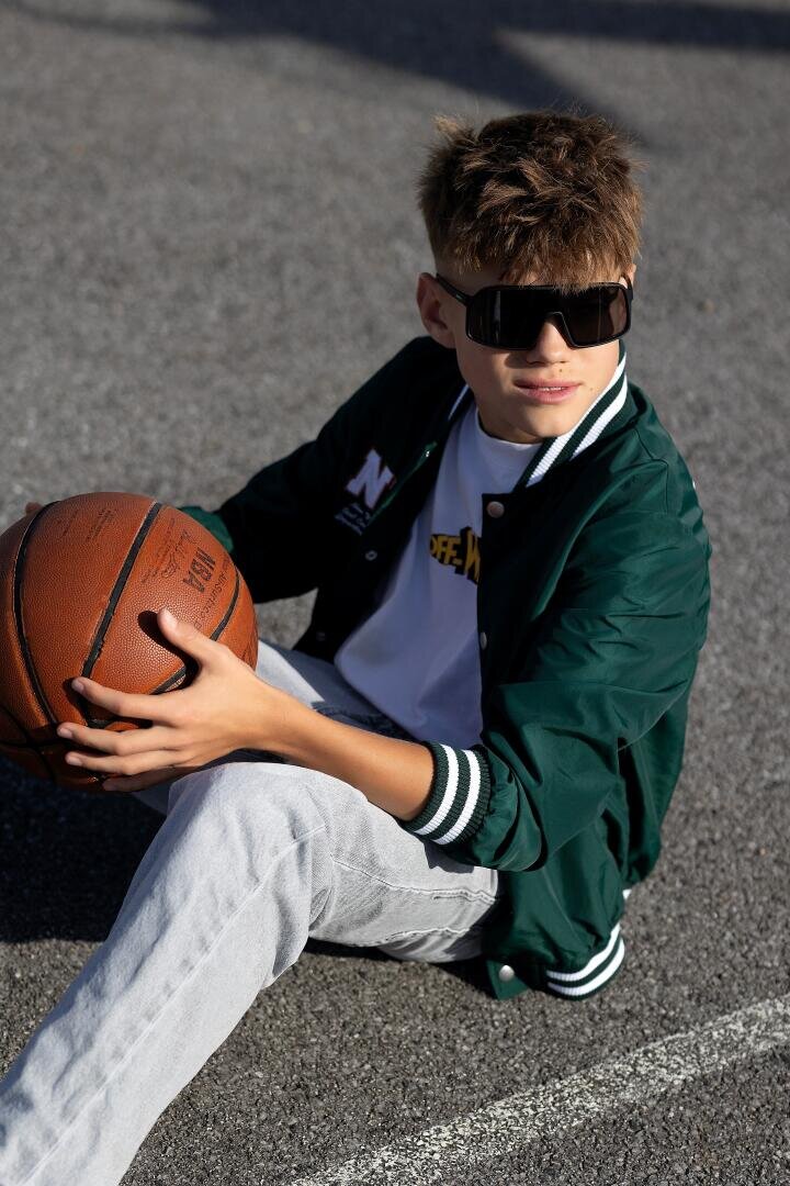 A boy with large sunglasses, a green jacket and light-colored jeans is sitting on an outdoor basketball court, holding a basketball and looking to the side with a relaxed expression.