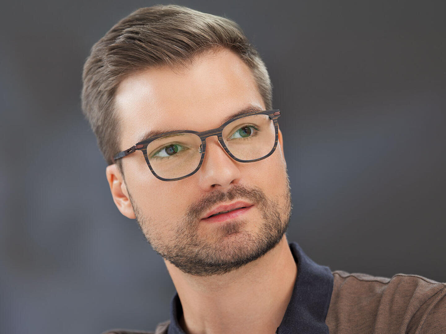 Ein junger Mann mit kurzen braunen Haaren, gestutztem Bart und dunkler Brille blickt leicht zur Seite vor einem dunklen, unscharfen Hintergrund. Er trägt ein legeres, dunkles Hemd mit Kragen.