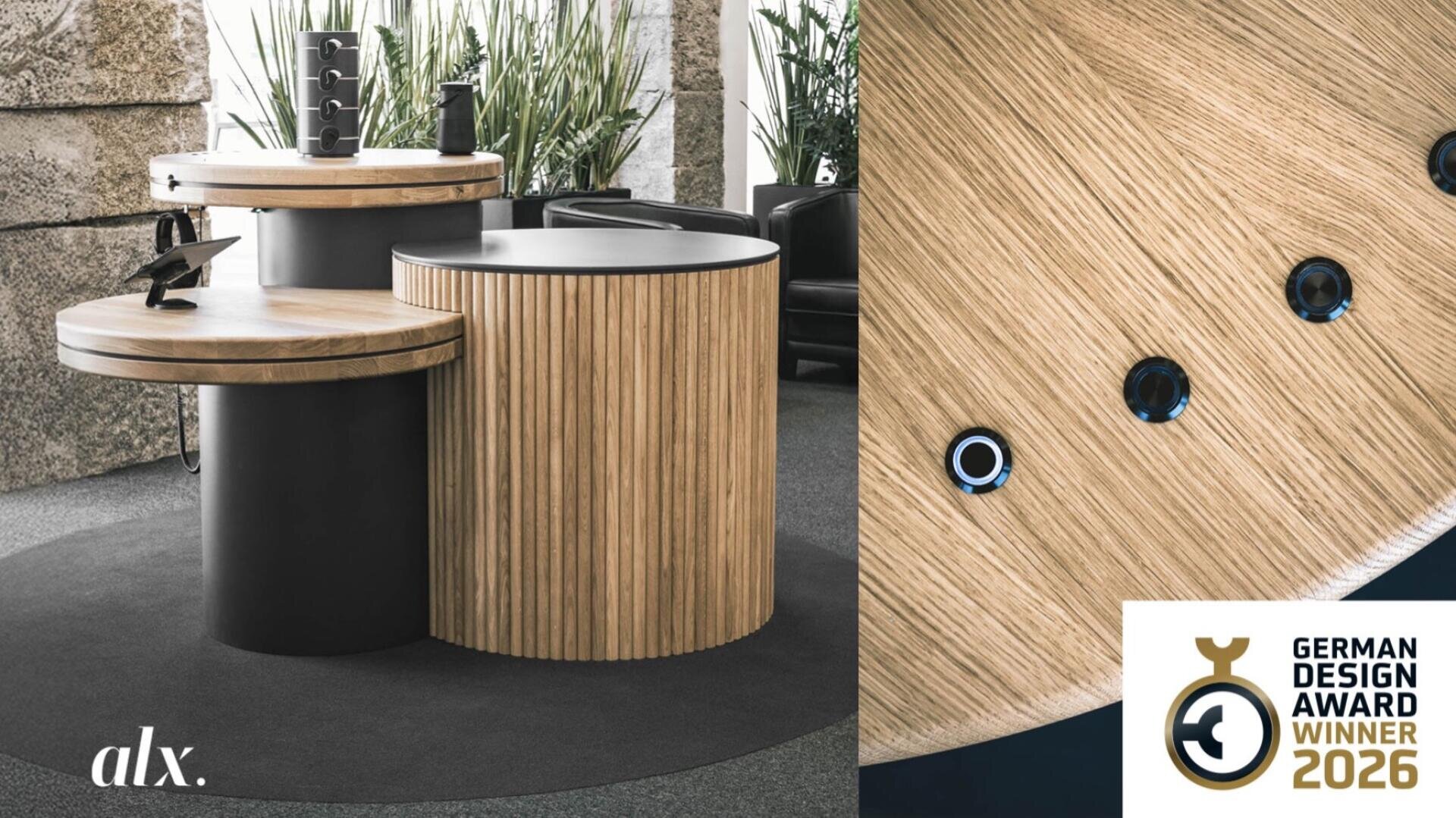 Ein moderner, runder Holztisch mit schwarzen Akzenten ist in einem Innenraum ausgestellt, umgeben von Pflanzen. In der rechten unteren Ecke ist eine Plakette des German Design Award Winner 2026 zu sehen, links unten ist alx. zu sehen.