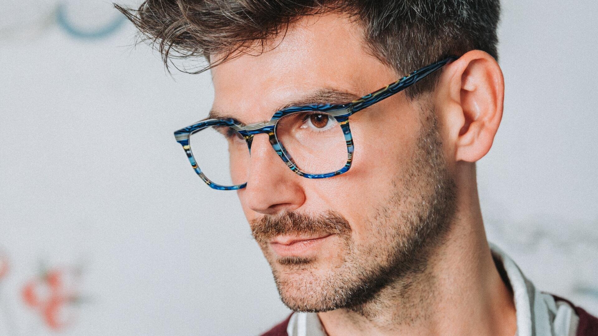 A man with short brown hair and a moustache wears colorful, blue-patterned glasses and looks slightly to the side against a light background.