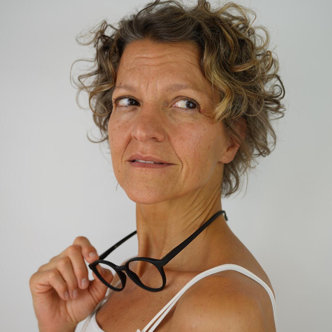 Eine Frau mittleren Alters mit lockigem kurzem Haar hält eine schwarze Brille in einer Hand. Sie trägt ein weißes Tank-Top und blickt leicht zur Seite mit einem nachdenklichen Ausdruck vor einem schlichten hellen Hintergrund.