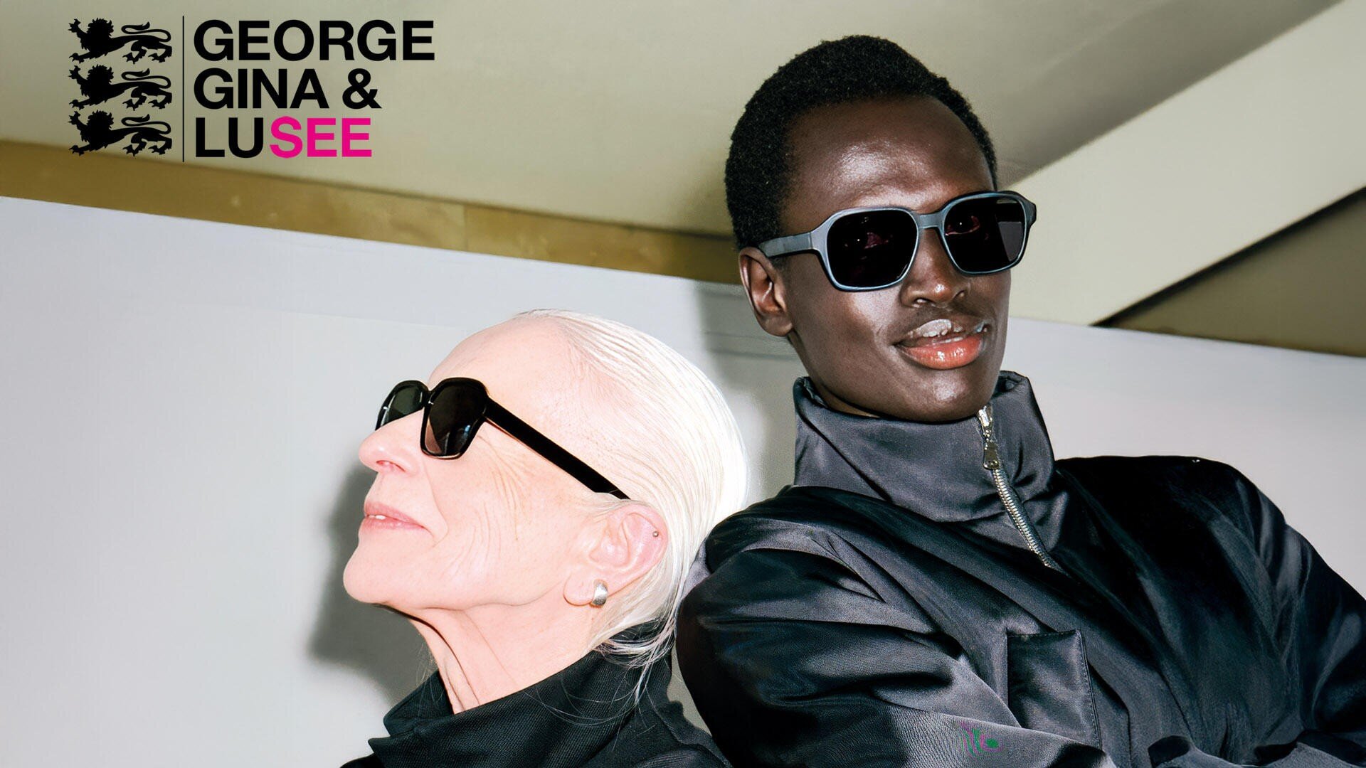 Zwei stilvolle Menschen mit dunklen Sonnenbrillen und schwarzen Jacken posieren selbstbewusst vor einem weißen Hintergrund. Das Logo von George Gina & Lucy erscheint in der oberen linken Ecke.