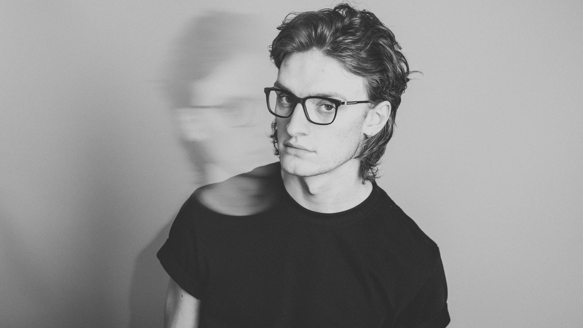 Eine junge Person mit Brille und welligem Haar trägt ein schwarzes T-Shirt und blickt ernst in die Kamera. Dahinter ist ein unscharfer Doppelbildeffekt vor einem schlichten Hintergrund zu sehen.