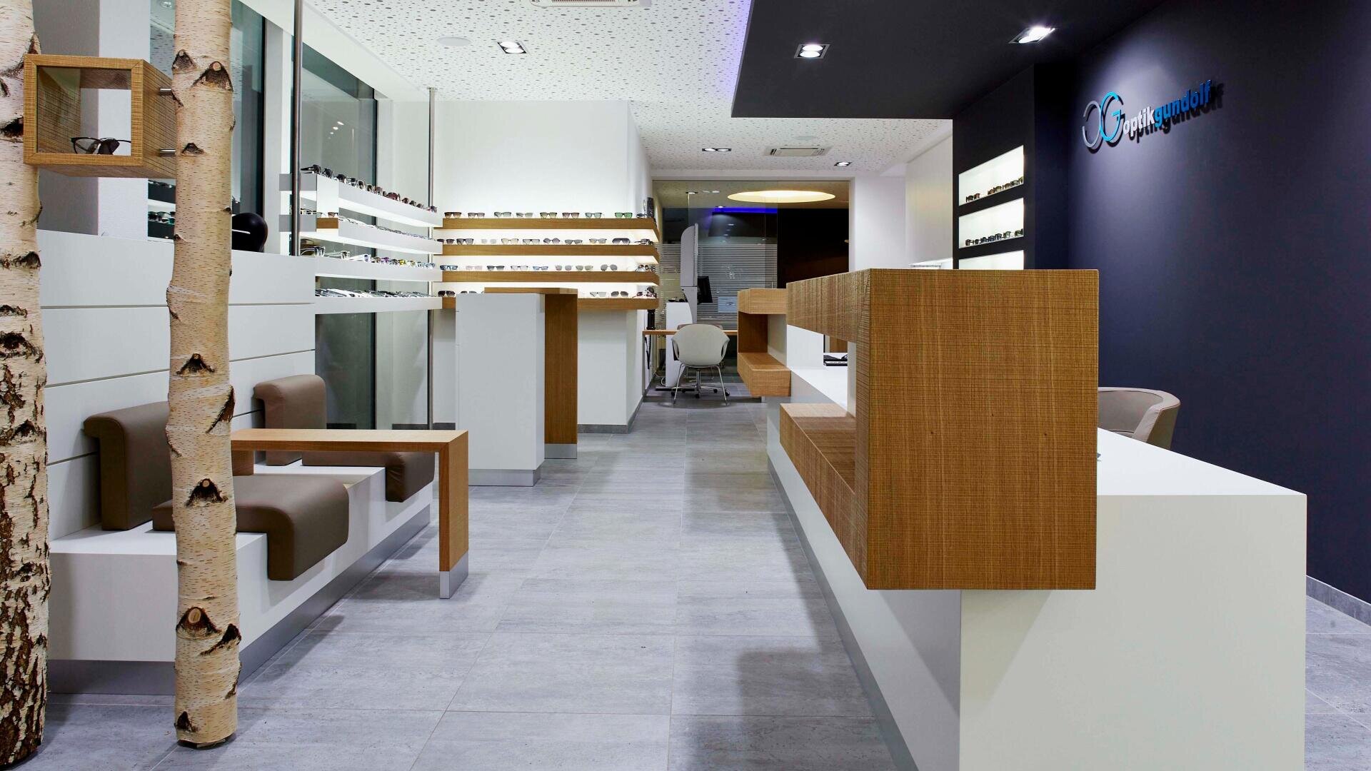 A modern optician's office with minimalist décor, wood and white furniture, shelves with glasses, a seating area and a reception desk. Birch trunks serve as decorative accents.