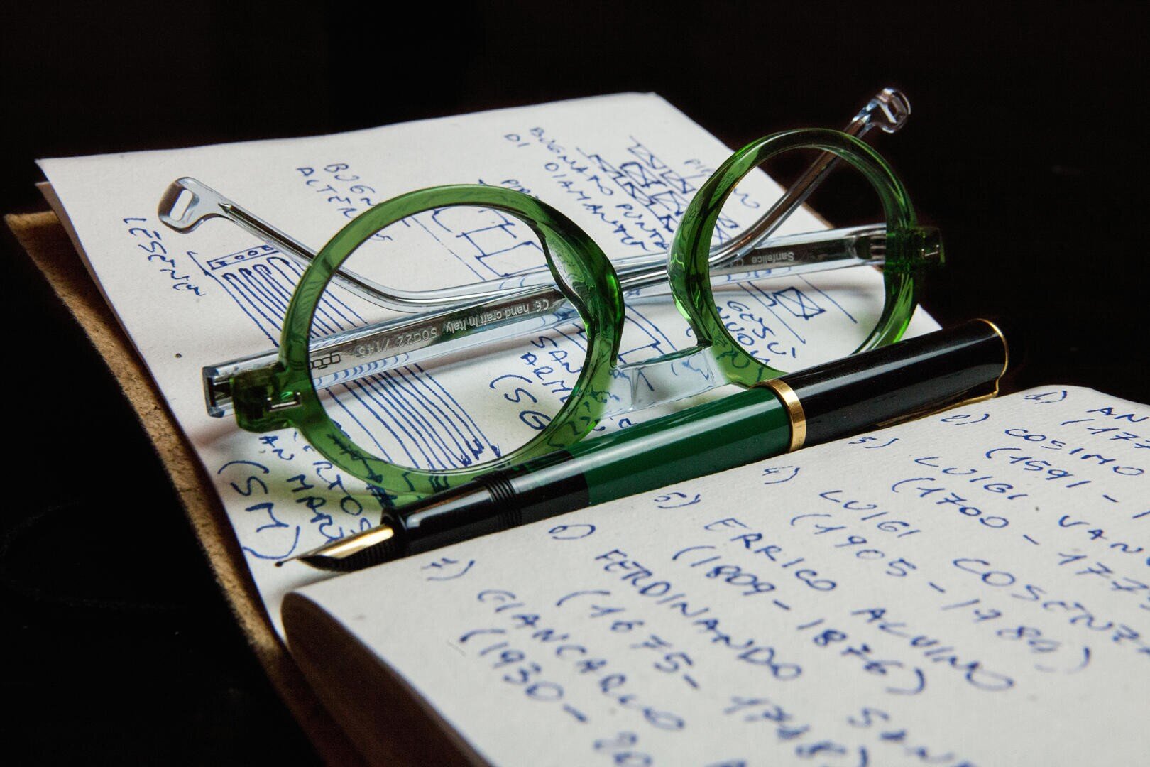 Eine grün umrandete Brille und ein grüner Füllfederhalter liegen auf einem aufgeschlagenen Notizbuch, das mit handschriftlichen Notizen in blauer Tinte gefüllt ist.