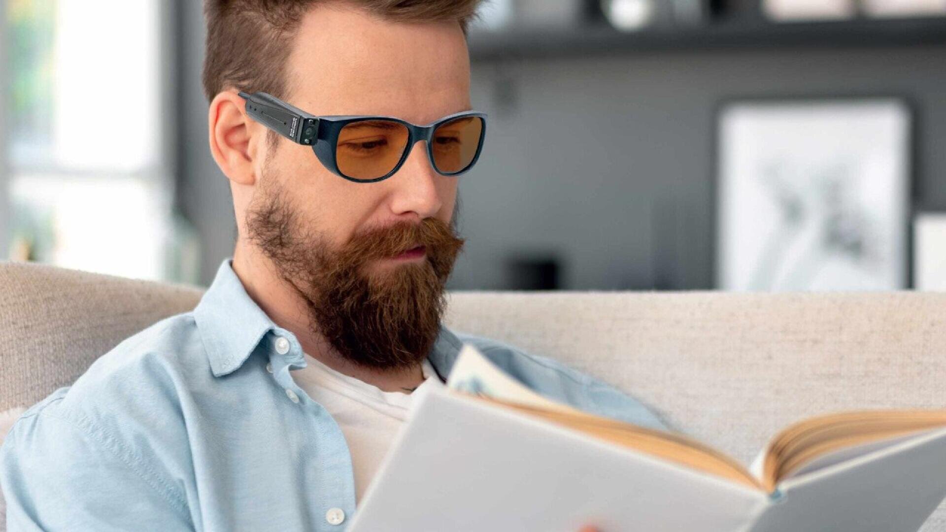 A man with a beard and glasses sits on a sofa, wearing a light blue shirt and reading a book. The background is softly blurred with shelves and framed pictures visible.