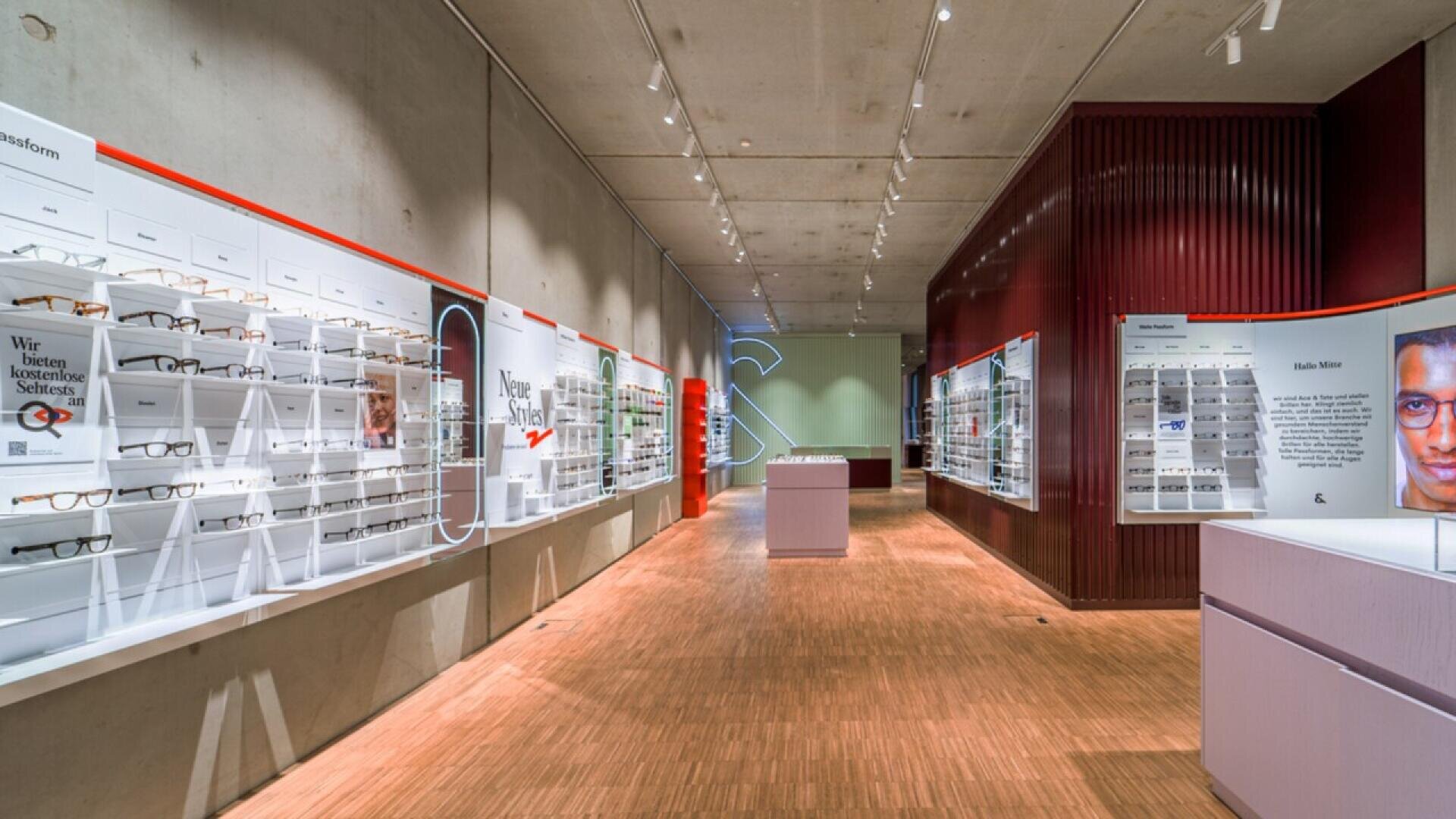A modern eyewear store with wooden floors and concrete walls displays numerous eyeglasses on white shelves along both sides of a well-lit hallway. A large image of a face is visible on the right wall.