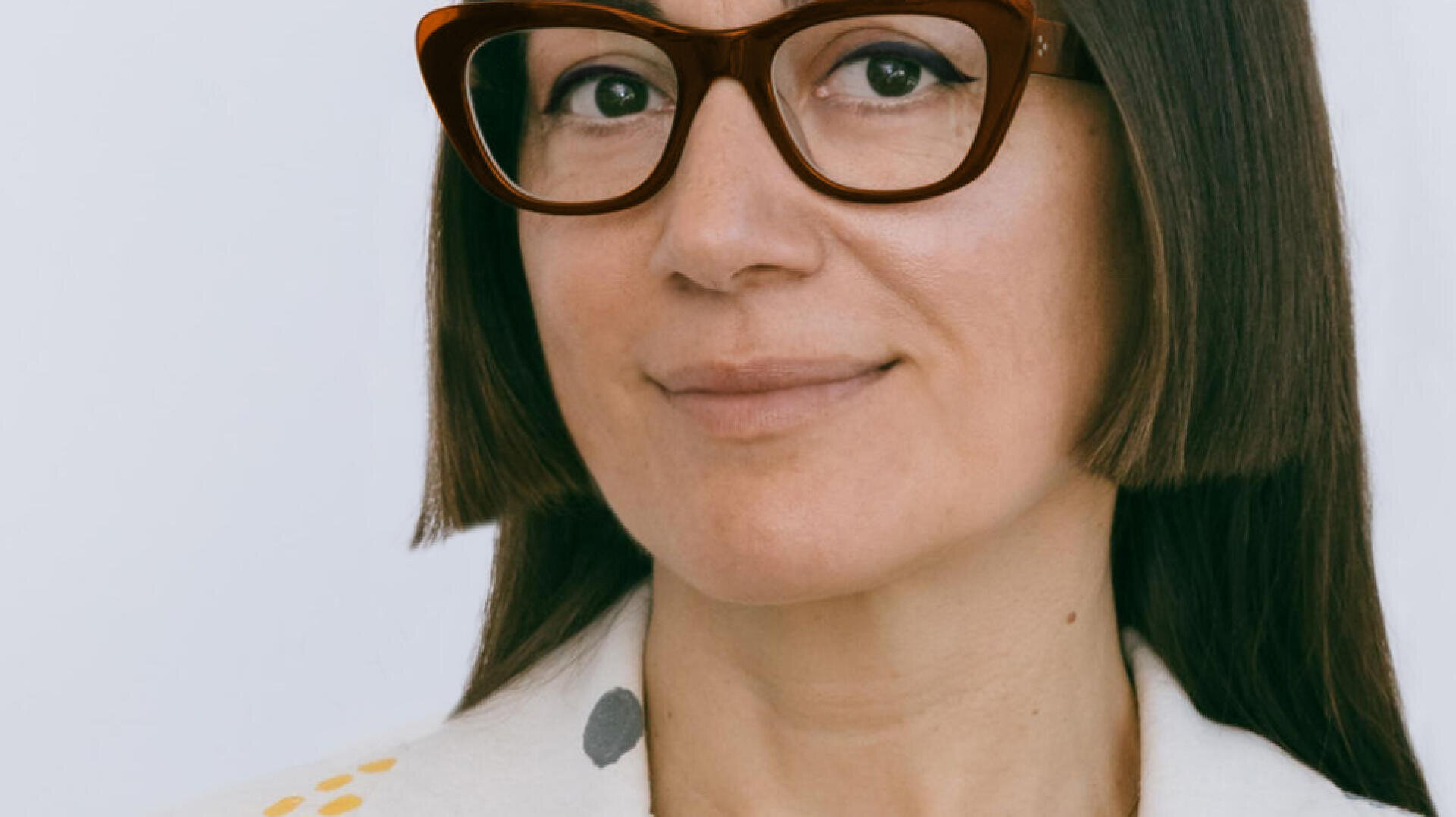 Eine Person mit glattem braunem Haar, einer übergroßen braunen Brille und einem weißen Oberteil mit gelben und grauen Punkten, die vor einem schlichten hellen Hintergrund leicht lächelt.