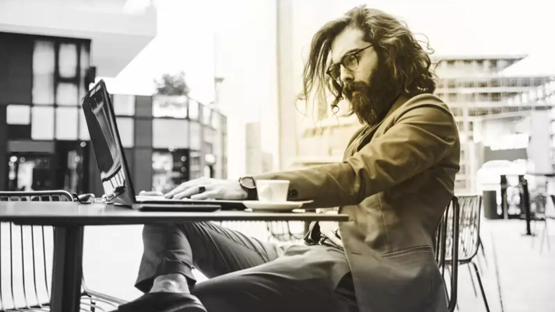 Ein Mann mit langen Haaren und Bart sitzt an einem Cafétisch im Freien und arbeitet an seinem Laptop. Er trägt Brille und Anzug. Daneben steht eine Tasse Kaffee. Im Hintergrund sind städtische Gebäude zu sehen.