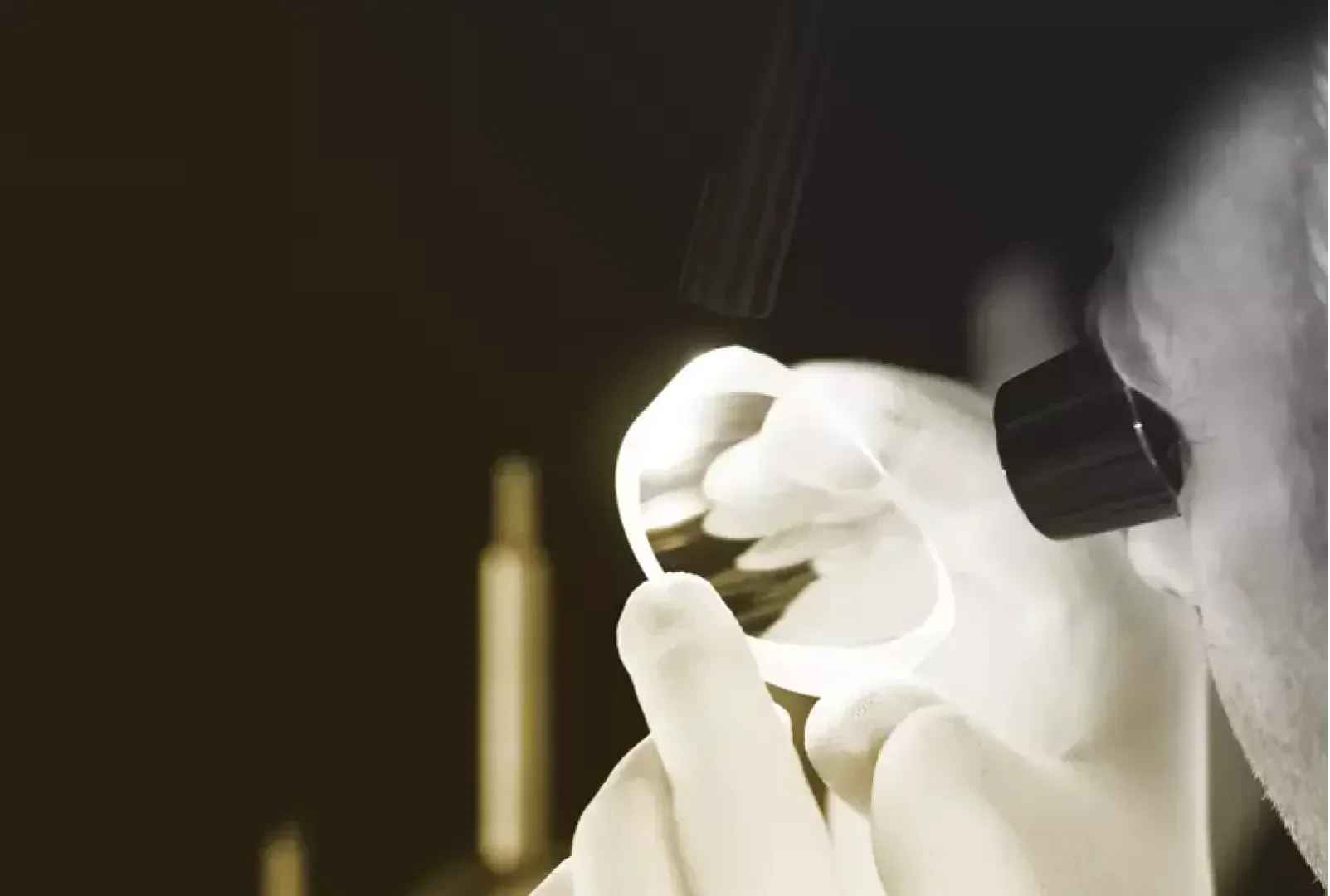 Eine Person mit weißen Handschuhen untersucht eine klare optische Linse unter einem Vergrößerungsgerät, wobei ein dunkler Hintergrund und eine fokussierte Beleuchtung die Linse hervorheben.