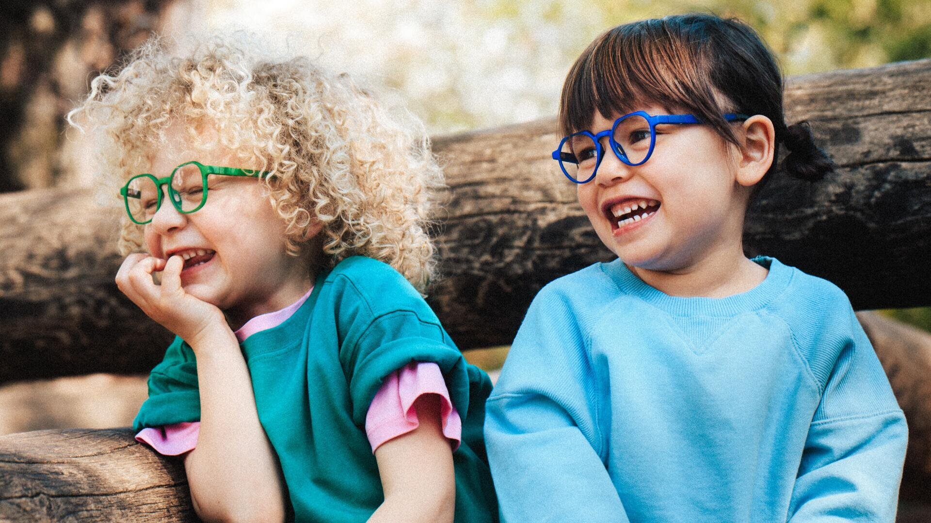 Zwei kleine Kinder mit bunten Brillen und einem Lächeln sitzen im Freien auf einem Holzzaun. Ein Kind hat lockiges blondes Haar und ein grünes Hemd, das andere hat glattes braunes Haar und trägt ein hellblaues Sweatshirt.