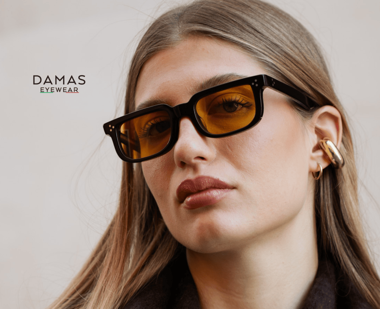 Eine Frau trägt eine rechteckige, schwarz gerahmte, gelb getönte Sonnenbrille und eine klobige goldene Ohrmanschette. Das Bild zeigt das Logo von DAMAS EYEWEAR auf einem neutralen Hintergrund.