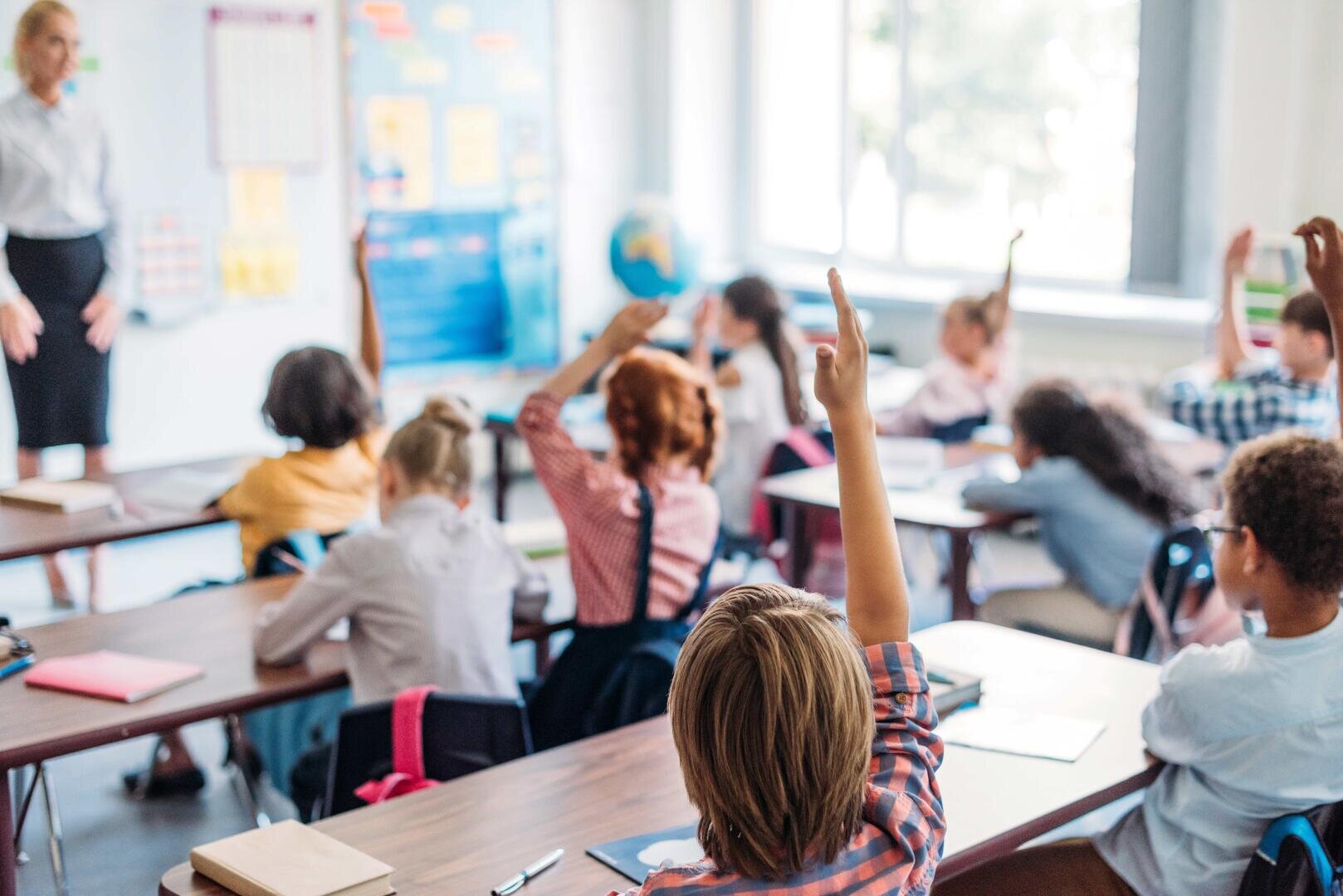 A classroom of young students sitting at desks, raising their hands to answer a question. A teacher stands at the front of the room, and sunlight comes through large windows in the background.