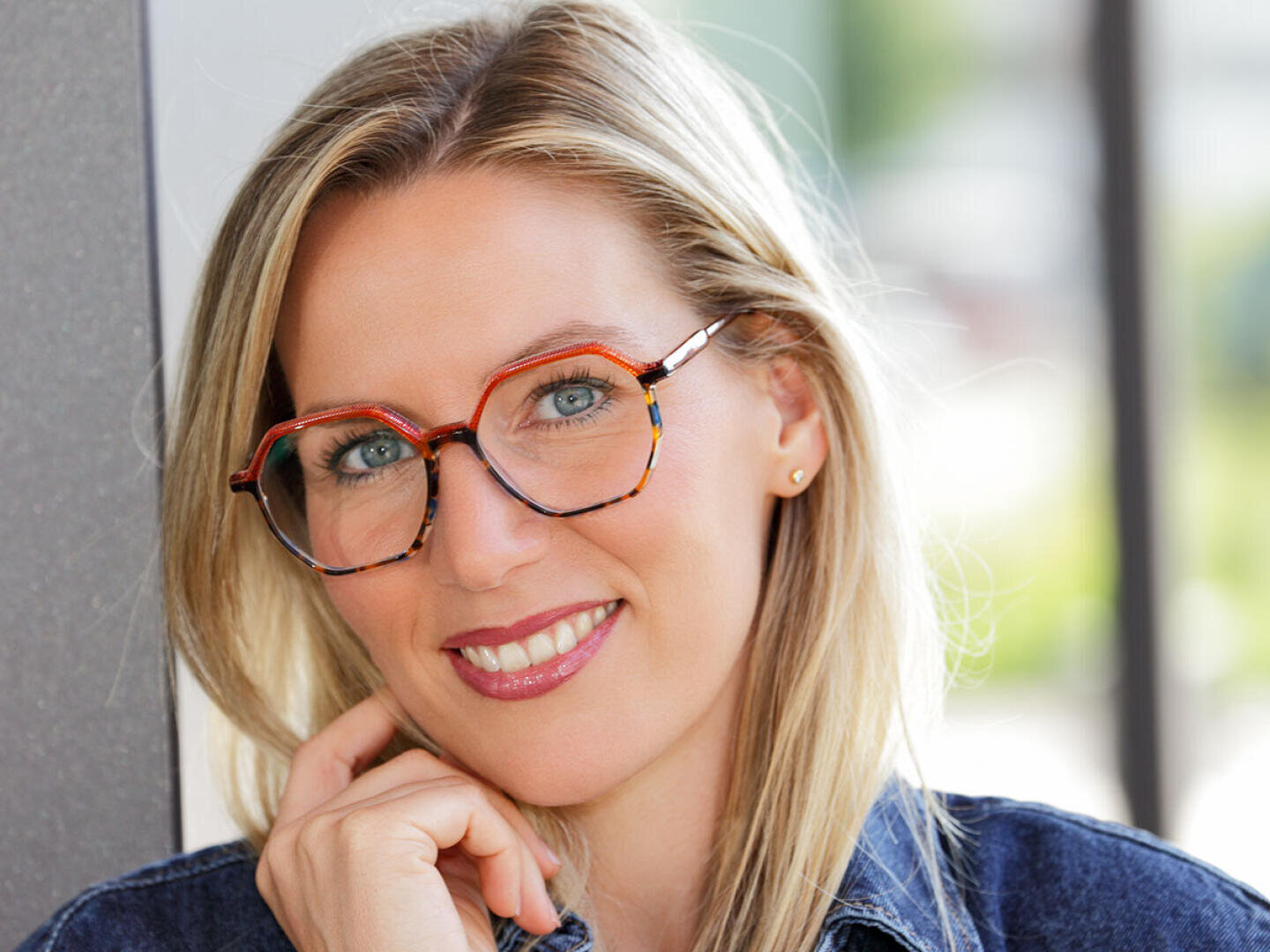 Eine lächelnde Frau mit langen blonden Haaren und hellblauen Augen trägt eine große, bunte Brille und eine Jeansjacke. Sie stützt ihr Kinn auf ihre Hand. Der Hintergrund ist leicht verschwommen.