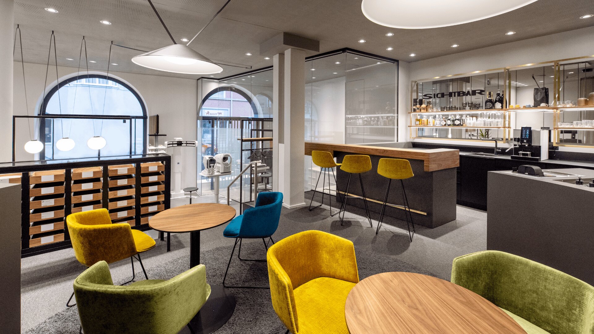 Moderner Lounge- und Barbereich mit farbenfrohen Samtstühlen, runden Holztischen, einem Bartresen mit Hockern, Regalen mit Flaschen und großen Bogenfenstern, die Tageslicht hereinlassen.