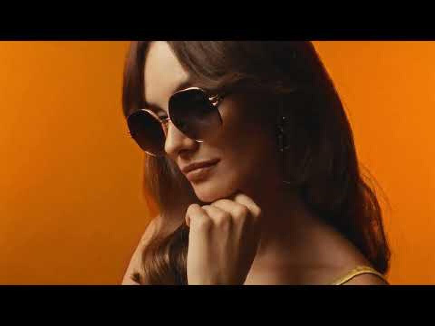 Eine Frau mit langen braunen Haaren und großer, runder Sonnenbrille posiert vor einem orangefarbenen Hintergrund, stützt ihr Kinn auf die Hand und schaut selbstbewusst zur Seite.