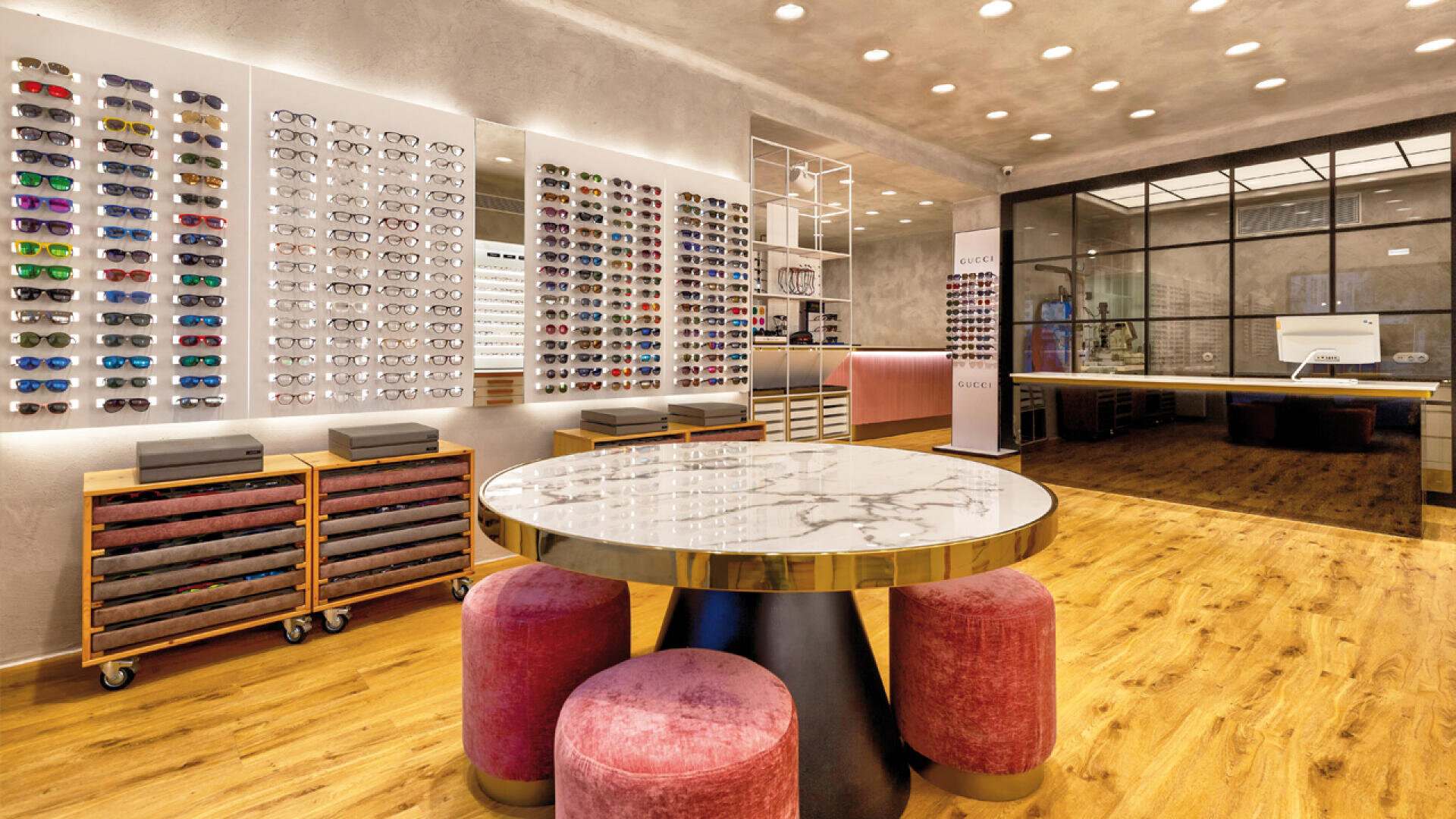 Modern eyewear store with wooden floors, a marble-topped round table surrounded by pink stools, wall displays of eyeglasses, drawers below, and a spacious, well-lit interior with glass partitions in the background.