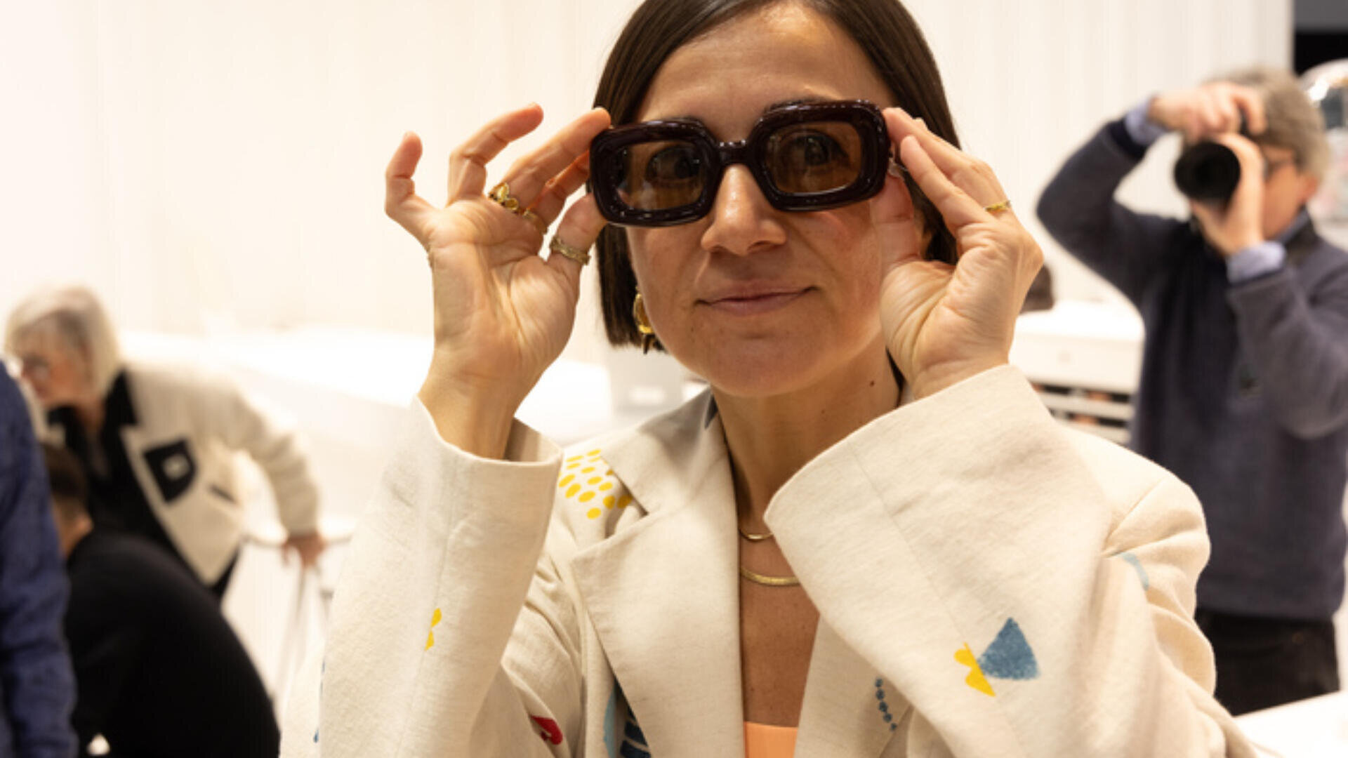 Eine Frau in einer hellen Jacke mit bunten Mustern hält sich eine übergroße dunkle Sonnenbrille vors Gesicht und lächelt. Im Hintergrund sind Menschen unscharf zu sehen, darunter auch jemand, der ein Foto macht.