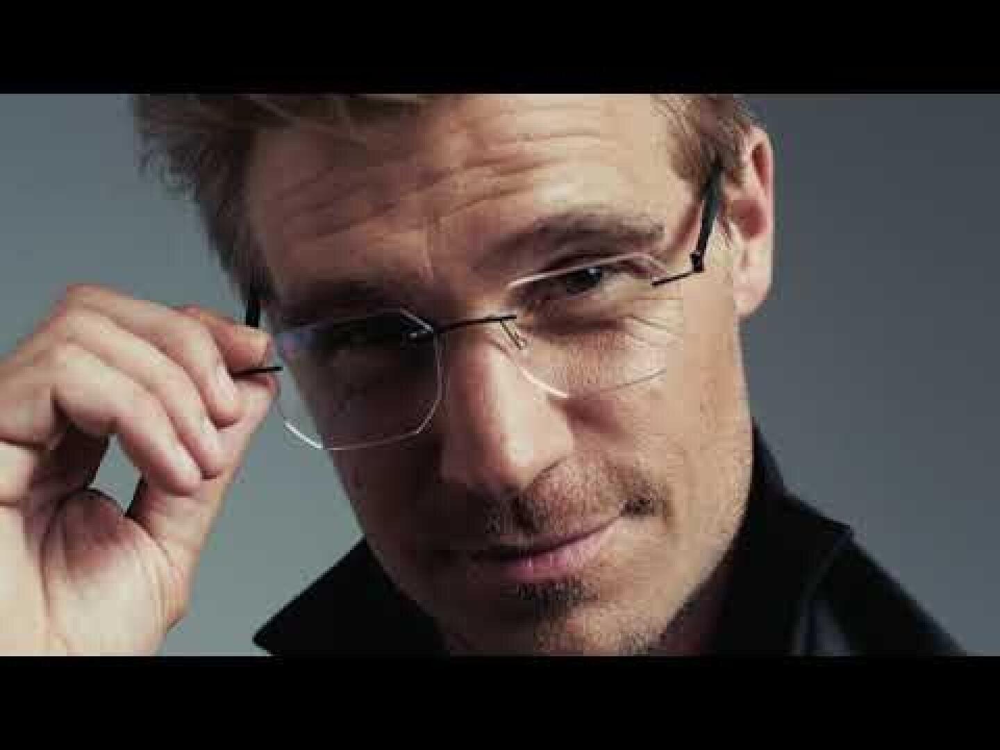 Ein Mann mit hellem Haar und Brille schaut selbstbewusst in die Kamera, während er mit einer Hand seine Brille zurechtrückt. Er ist mit einer dunklen Jacke vor einem neutralen Hintergrund bekleidet.