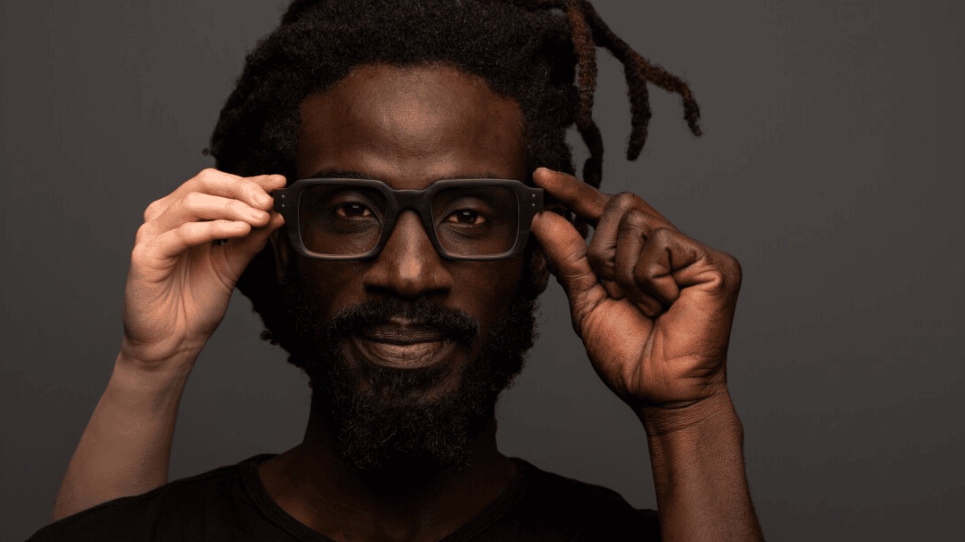 A man with a beard and dreadlocks smiles gently while adjusting black glasses. A pair of light-skinned hands from behind holds the glasses, helping to place them on his face against a neutral background.