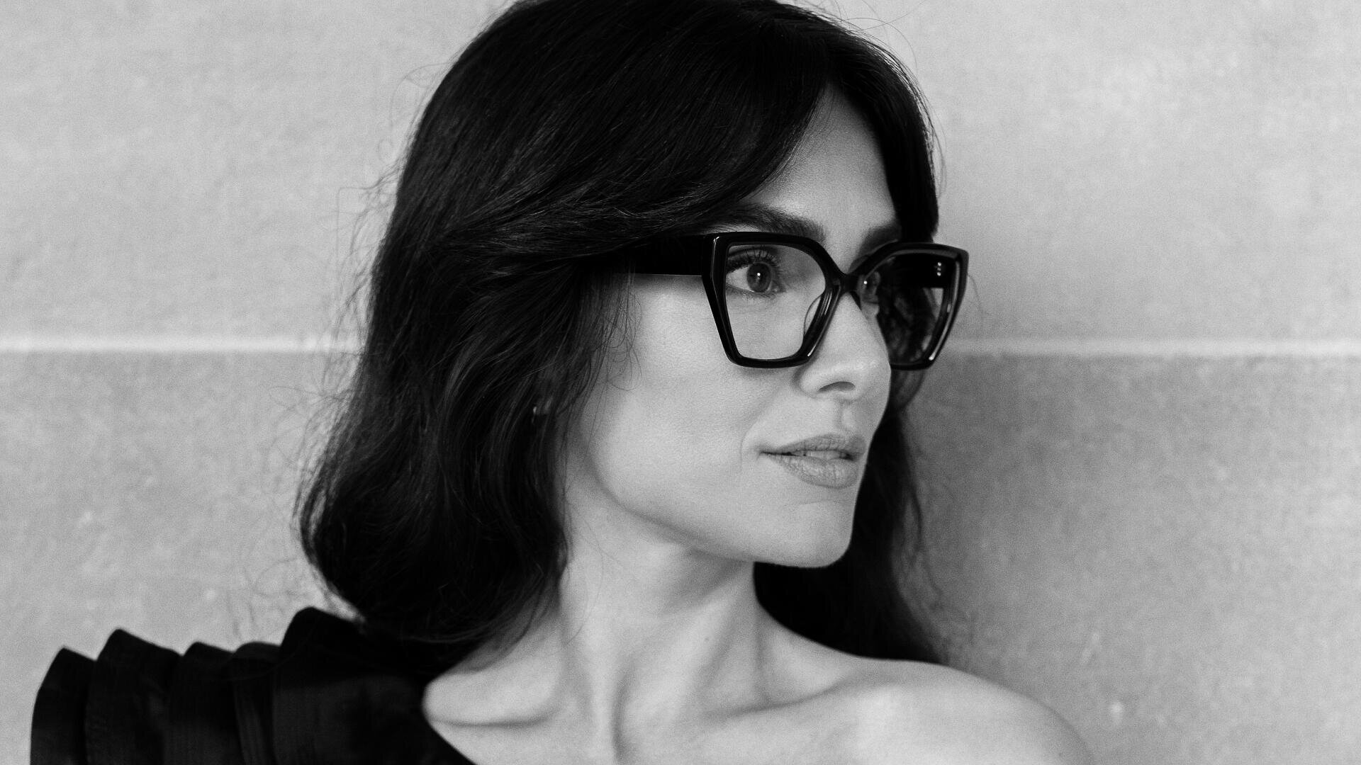 Black-and-white photo of a woman with long dark hair wearing large square glasses and an off-shoulder top, looking to the side in front of a plain textured wall.