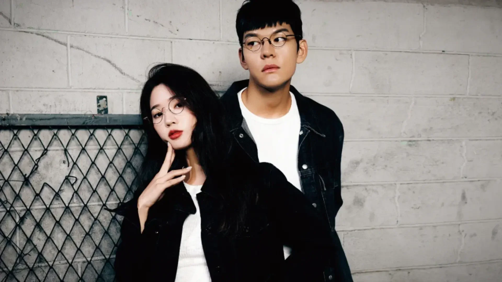 A young woman and man, both wearing glasses, black jackets, and white shirts, stand against a gray brick wall. The woman poses with one hand near her face; the man stands slightly behind her, looking to the side.