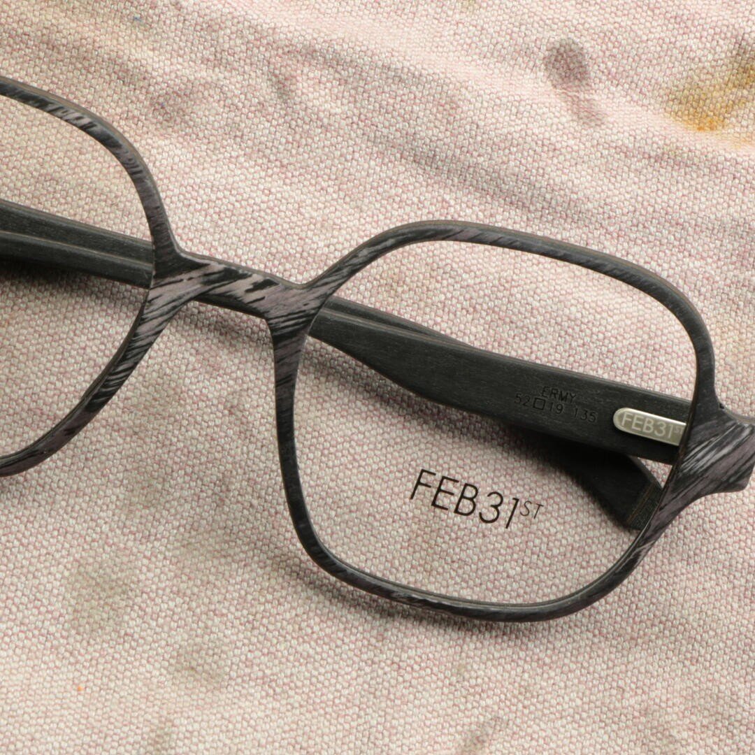 Close-up of a dark wooden spectacle frame resting on a textured beige fabric. The text "FEB31st." is engraved on the inside of the temples and lenses.